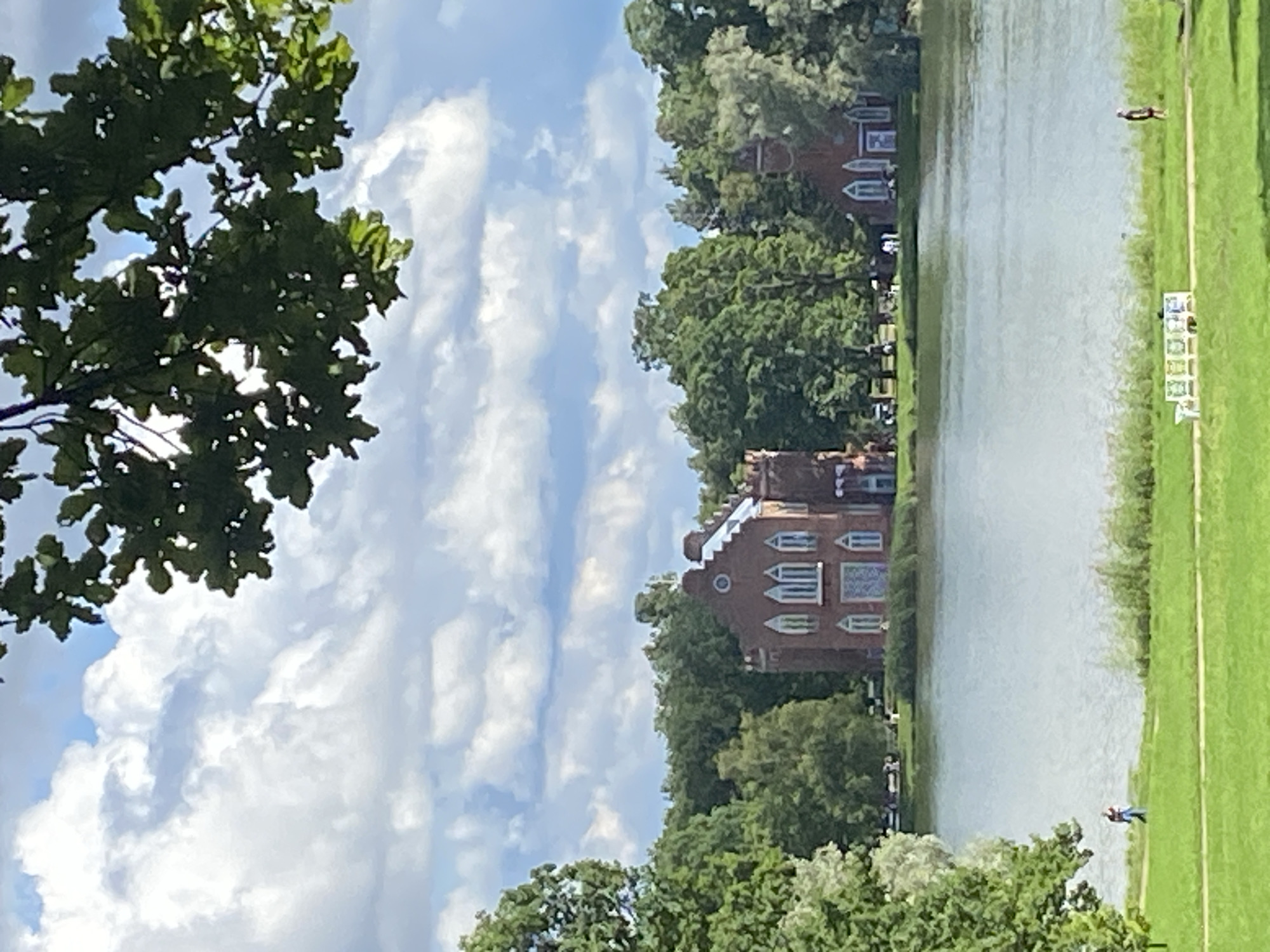 This is a photograph taken from a distance, showing a large brick building with arched windows on the shore of a lake. The building appears to be a historical structure, possibly a palace or a museum, situated within a park or estate. Several trees surround the building and are visible in the background, forming a dense green forest. The sky is partly cloudy, with white clouds scattered across a blue sky. In the foreground, there is a grassy area with a path and some benches. Two individuals are visible walking on the grass near the lake. The overall impression is one of a serene and picturesque landscape. Based on the architectural style and setting, this scene is likely located in the city of Pushkin, Russia, possibly at the Catherine Palace or its surrounding park.
