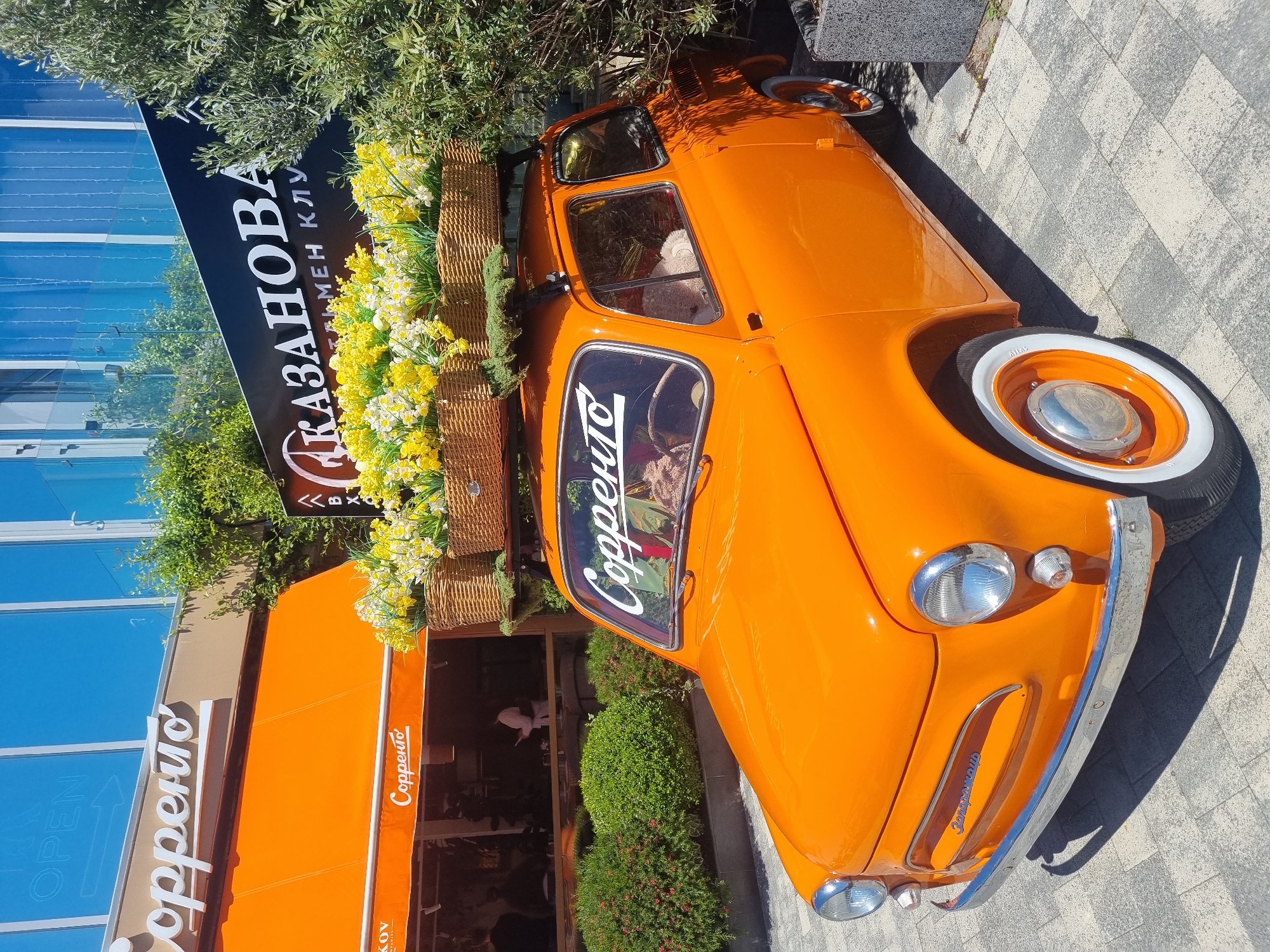 The image shows an orange vintage car, likely a ZAZ-965, parked on a sidewalk in what appears to be a commercial area. The car has white-wall tires and chrome trim, and its roof is adorned with flower baskets filled with yellow and white blossoms. The car seems to be used as a decorative element or advertisement for a restaurant called "Copperito". The restaurant is located next to a building. Signage next to the car bears Cyrillic text that includes the name "Казанова" (Kazanova). The location is Sochi, Russia.