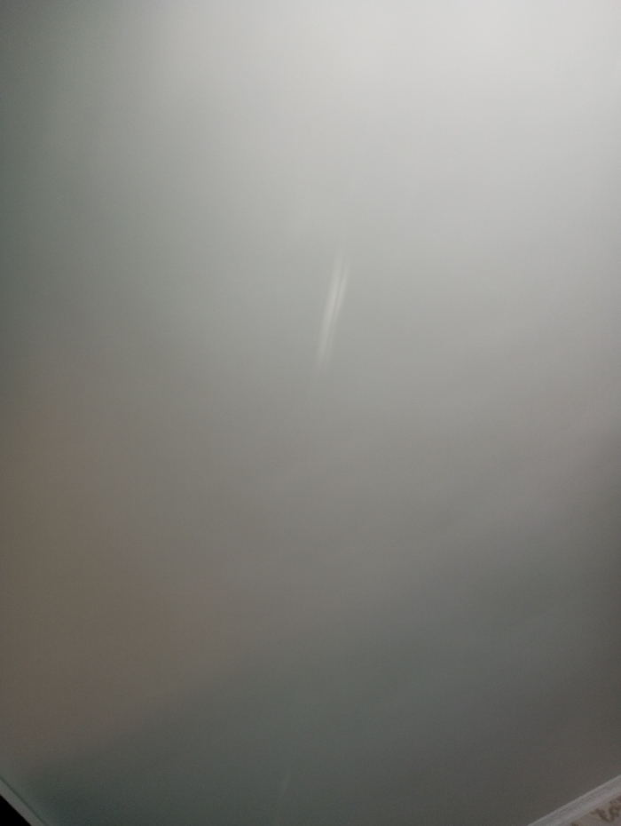 The image displays a uniform, light gray interior surface, likely a wall or ceiling, located within the city of Яромаска, Russia. A distinct, elongated, bright white vertical streak is centrally positioned in the upper-right quadrant of the frame, approximately 10-15% from the right edge and extending downwards. The surface exhibits subtle variations in tone, with diffuse lighting originating from an unseen source, causing a slight gradient from lighter values in the upper-right to darker values in the lower-left. In the extreme bottom-right corner, a partial view of a patterned, light-colored edge or surface element is visible, possibly indicating a floor or a baseboard. No individuals, additional specific objects, or discernible actions or interactions are present within the frame.