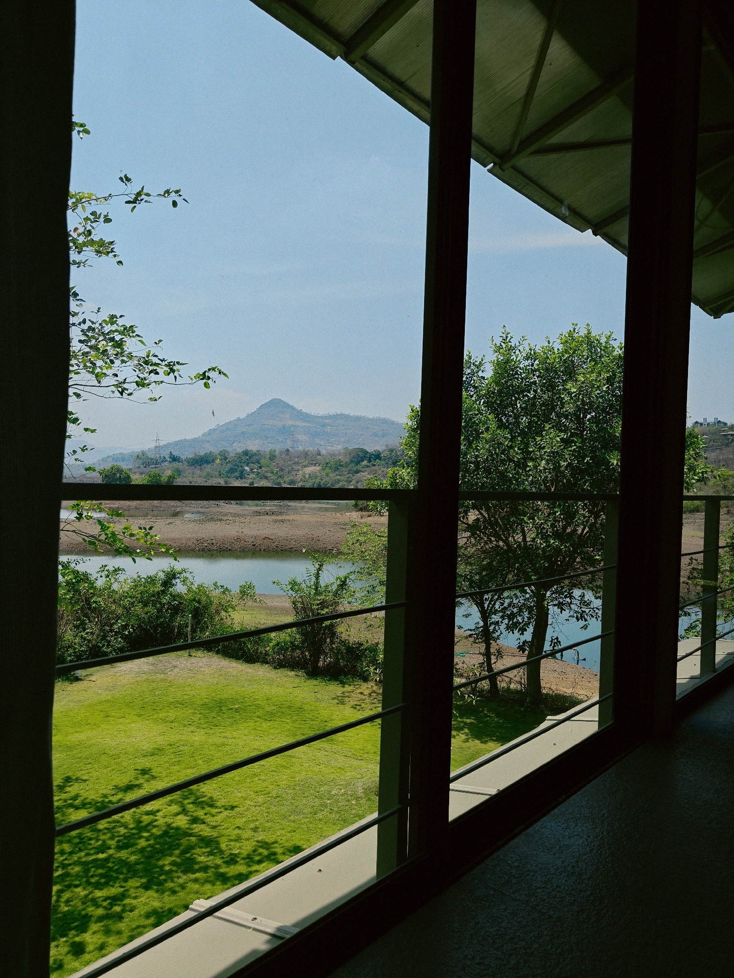 View from inside a building in Kalote Mokashi, India, showing a landscape with a body of water, grassy area, and a hill in the distance. The view is framed by architectural elements like pillars and a roof overhang. A railing is visible outside the building. The water level appears low, exposing a muddy shoreline. Trees and shrubs are interspersed across the landscape.