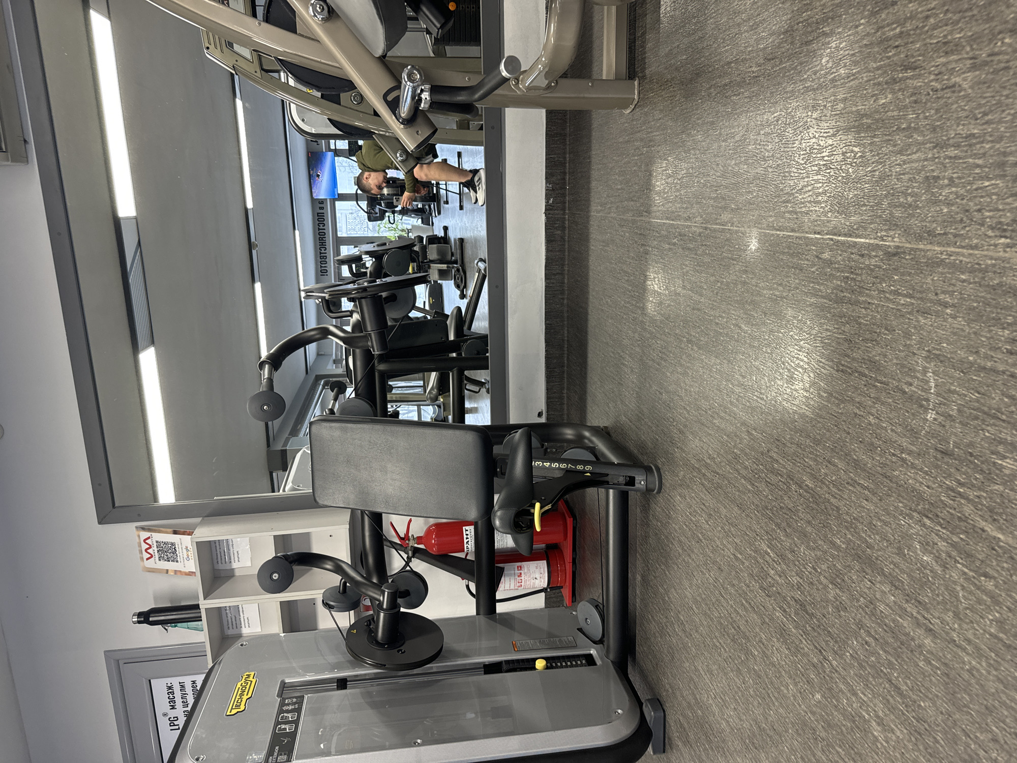 The image is an indoor view of a gym, likely located in Varna, Bulgaria, considering the signage in Bulgarian. Gym equipment dominates the foreground, particularly a black and gray weight machine with a padded seat. A red fire extinguisher is attached to the machine. The floor is a gray wood-grain pattern. A large mirror is mounted on the wall, reflecting a broader view of the gym. A person is using a weight machine in the reflection, clad in a green top and shorts. A white shelving unit with various items is visible next to the mirror. Signage with text, a QR code, and a Google logo is attached to the shelf.