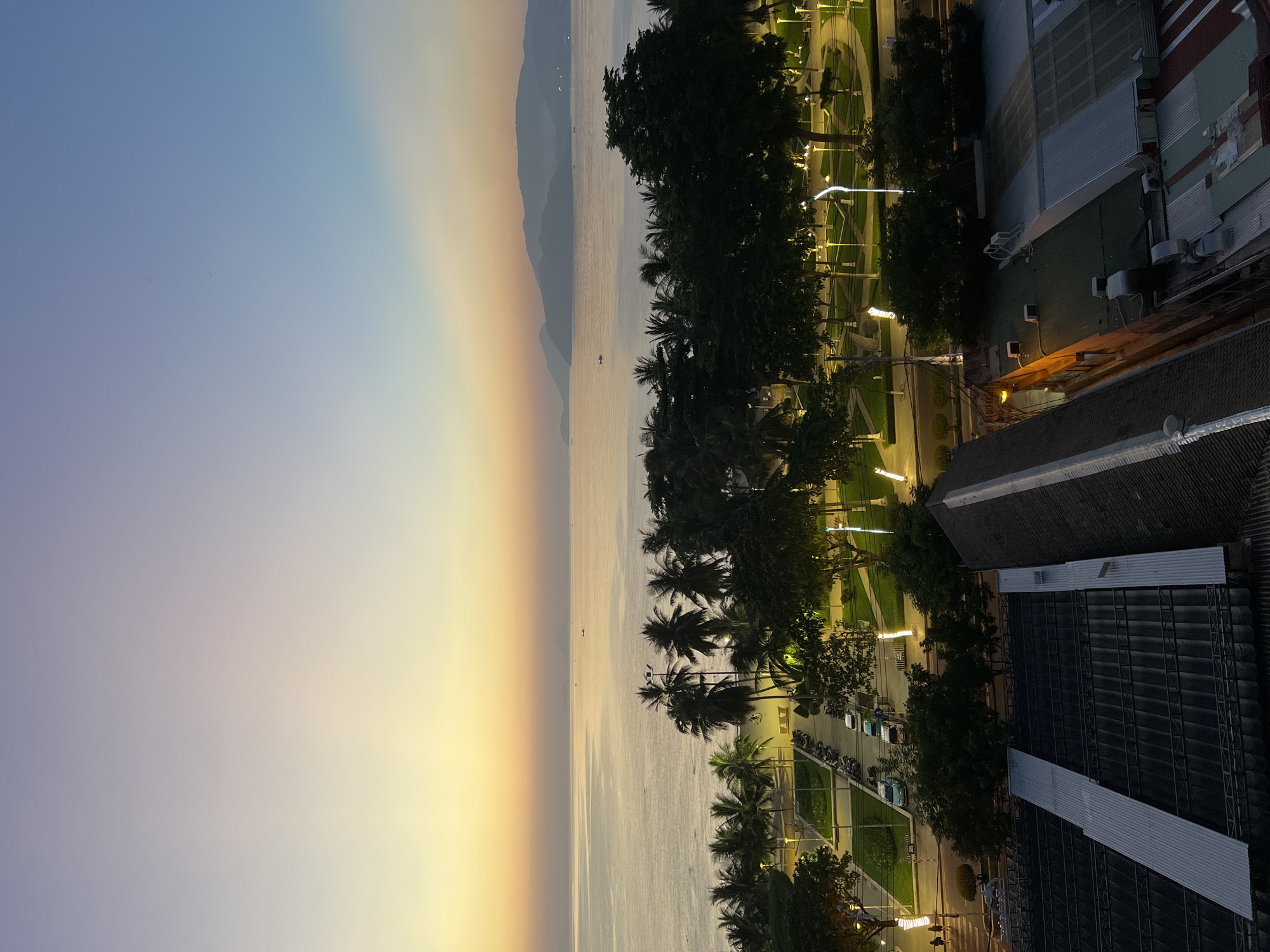 This image depicts a panoramic view of a coastal city at dawn, likely Nha Trang, Vietnam, given the presence of palm trees and the architecture. The sky transitions from a soft lavender and pale yellow near the horizon to a clear blue overhead. The calm sea stretches out, reflecting the dawn light, with distant hazy mountains visible on the horizon. Several small boats are scattered on the water.
In the foreground, a well-maintained park area with manicured green lawns and winding pathways is illuminated by decorative streetlights. A line of tall palm trees borders the park, creating a tropical ambiance. Adjacent to the park is a street with parked motorcycles and a few cars, suggesting a typical urban scene.
Further down, the rooftops of several buildings are visible. One prominent structure features a dark, sloped roof with metal sheeting. The buildings appear to be a mix of commercial and residential properties, with visible air conditioning units and other rooftop fixtures. The overall scene captures the tranquil transition from night to day in a coastal Vietnamese city.