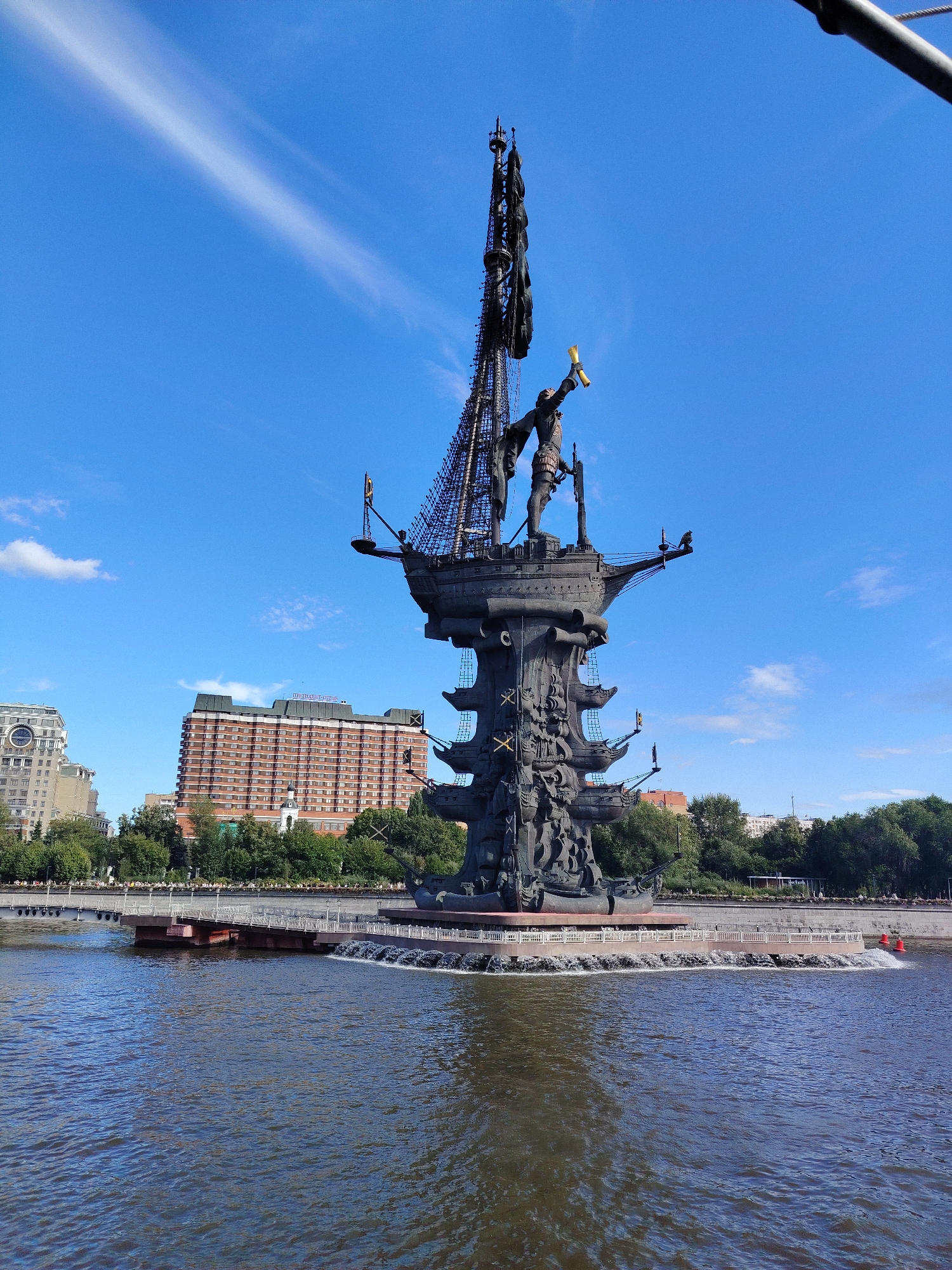 A large bronze statue of Peter the Great stands on a boat-shaped pedestal in a body of water. The statue depicts Peter the Great holding a scroll and pointing. The pedestal is ornate, with relief sculptures and multiple levels. In the background, there are trees, buildings, and a blue sky with some clouds. The body of water is a river or canal, with ripples on the surface. The statue is located on an island in the Moskva River in Moscow, Russia.