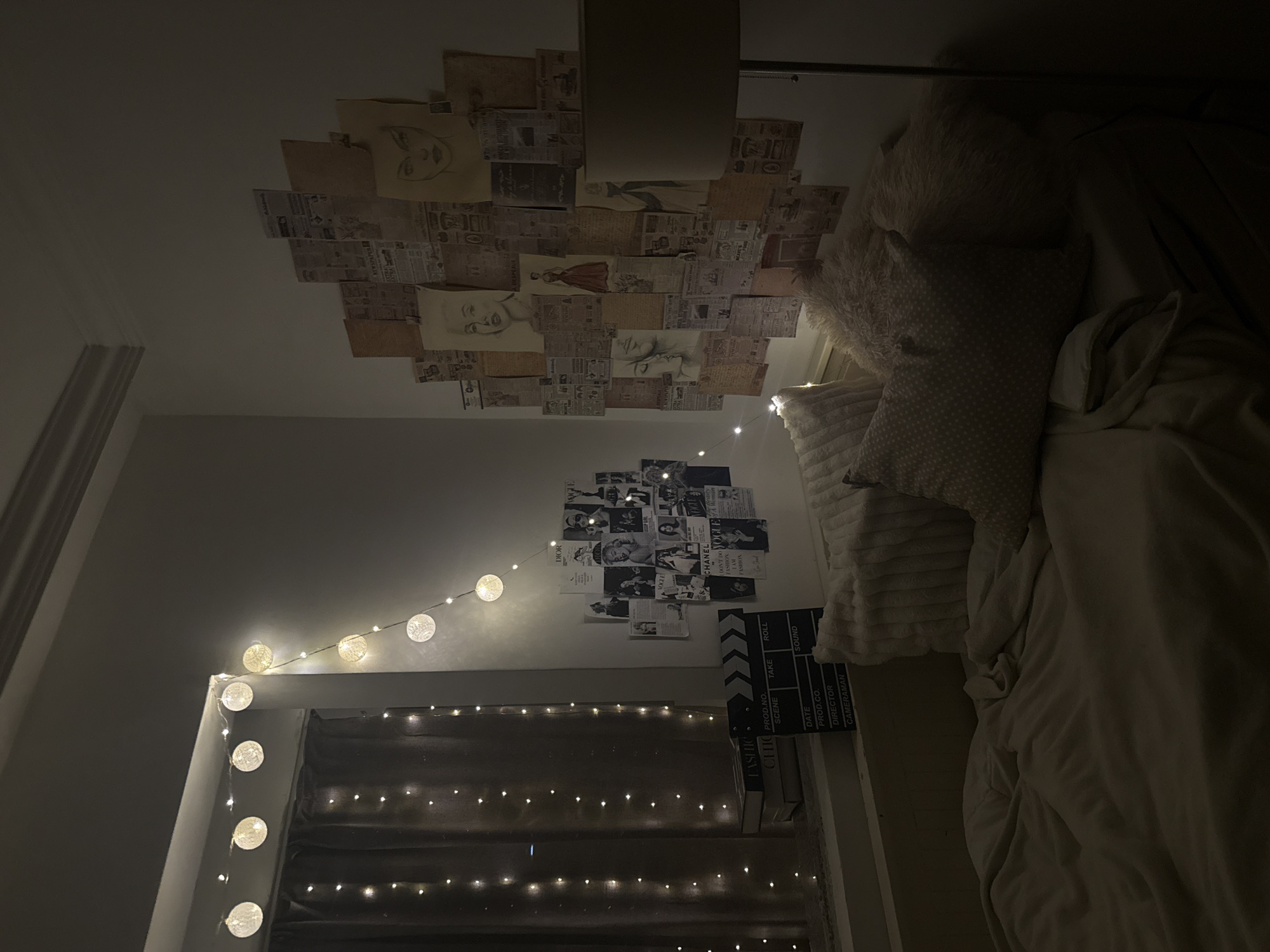 The image shows a bedroom interior, likely at night given the dim lighting. The ceiling is decorated with a collage of newspapers and portrait drawings. On the wall, there's another collage with black and white magazine covers. String lights are draped along the window frame and the wall. A film slate leans against the wall, near books, and pillows and bedding are partially visible on the right side of the frame. The location is not identifiable as Sofia, Bulgaria based on the image alone.