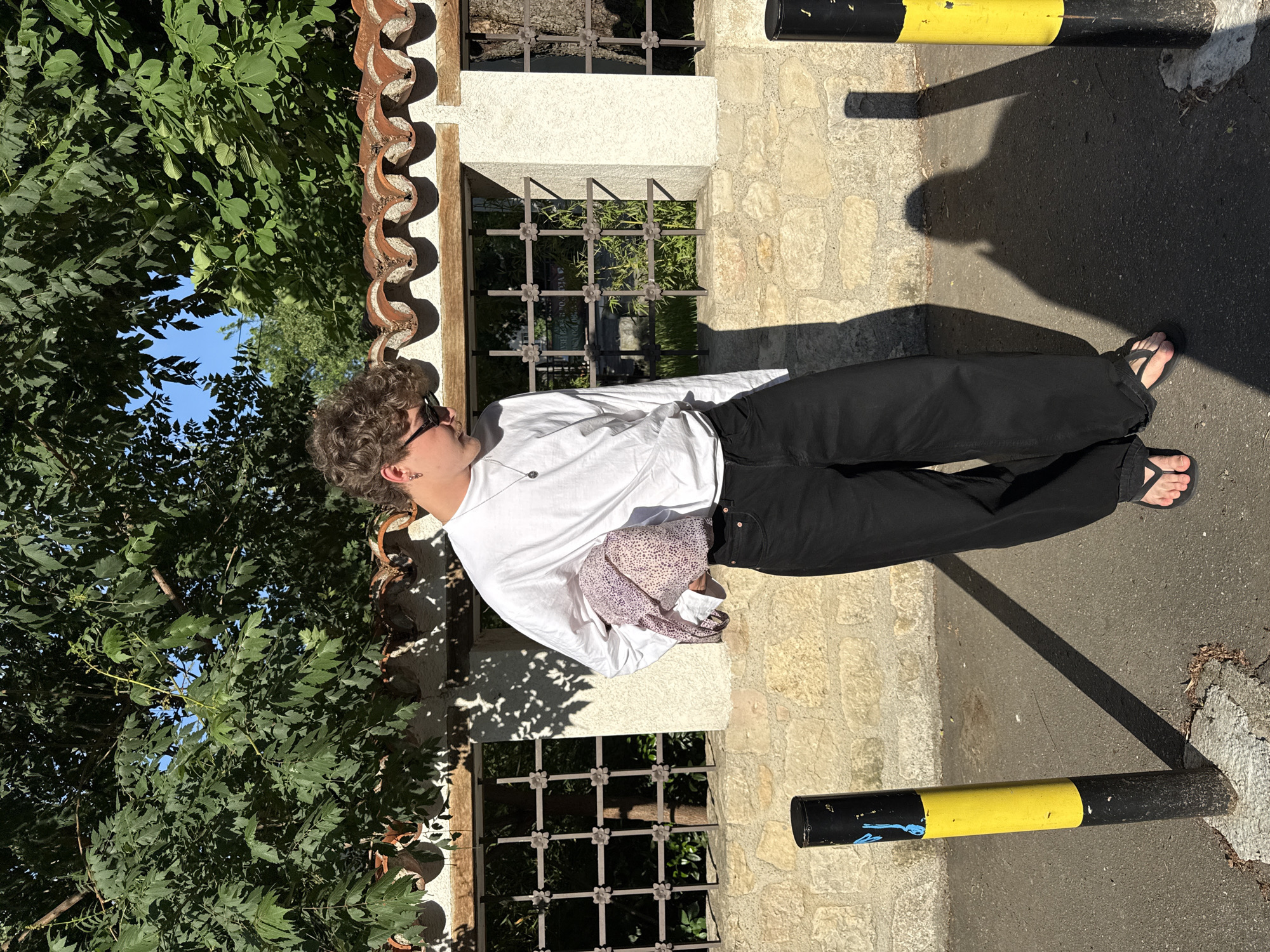 A person stands beside a stone wall with iron-grated windows, adjacent to a tree and a black and yellow bollard on a paved surface. The individual is wearing black pants, a white shirt, and flip-flops. The setting is illuminated by sunlight, casting shadows. The location is likely Belgrade, Serbia.
