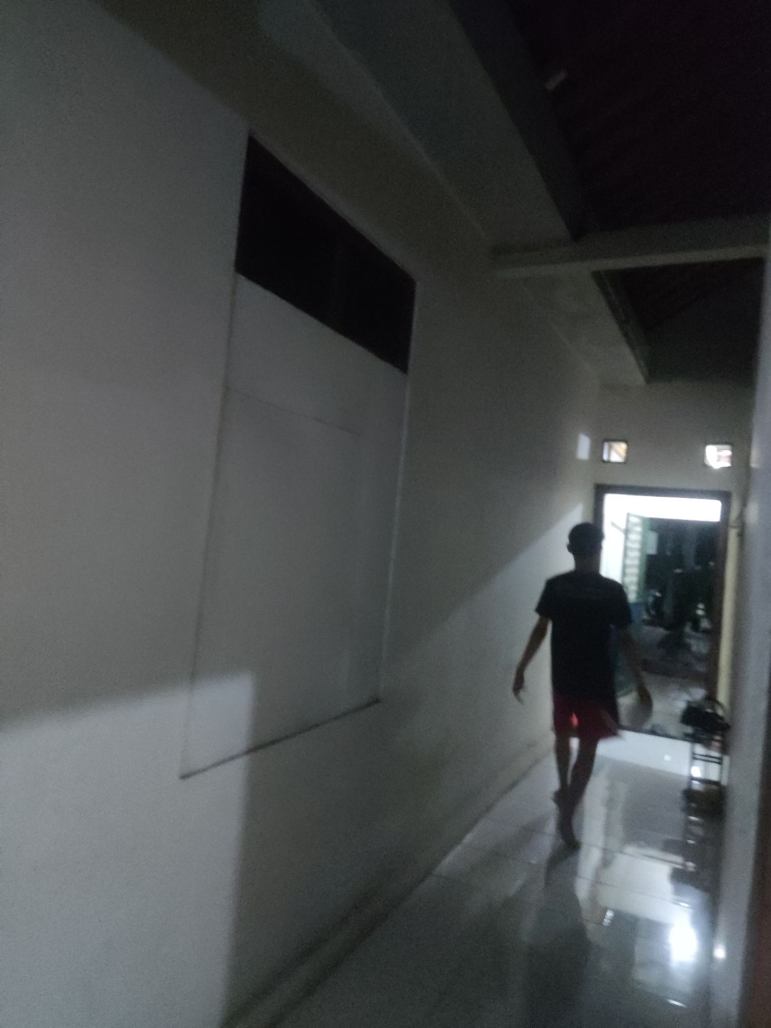 The image depicts a dimly lit hallway with a person walking away from the camera. The hallway is lined with plain walls and has a tiled floor that reflects the light. There are small square windows on the wall, and a doorway at the end of the hallway with light emanating from it. The person is wearing a dark t-shirt and red shorts. The scene appears to be in Trihanggo, Indonesia, likely within a residential or commercial building due to the architectural style. The overall atmosphere is subdued and quiet.