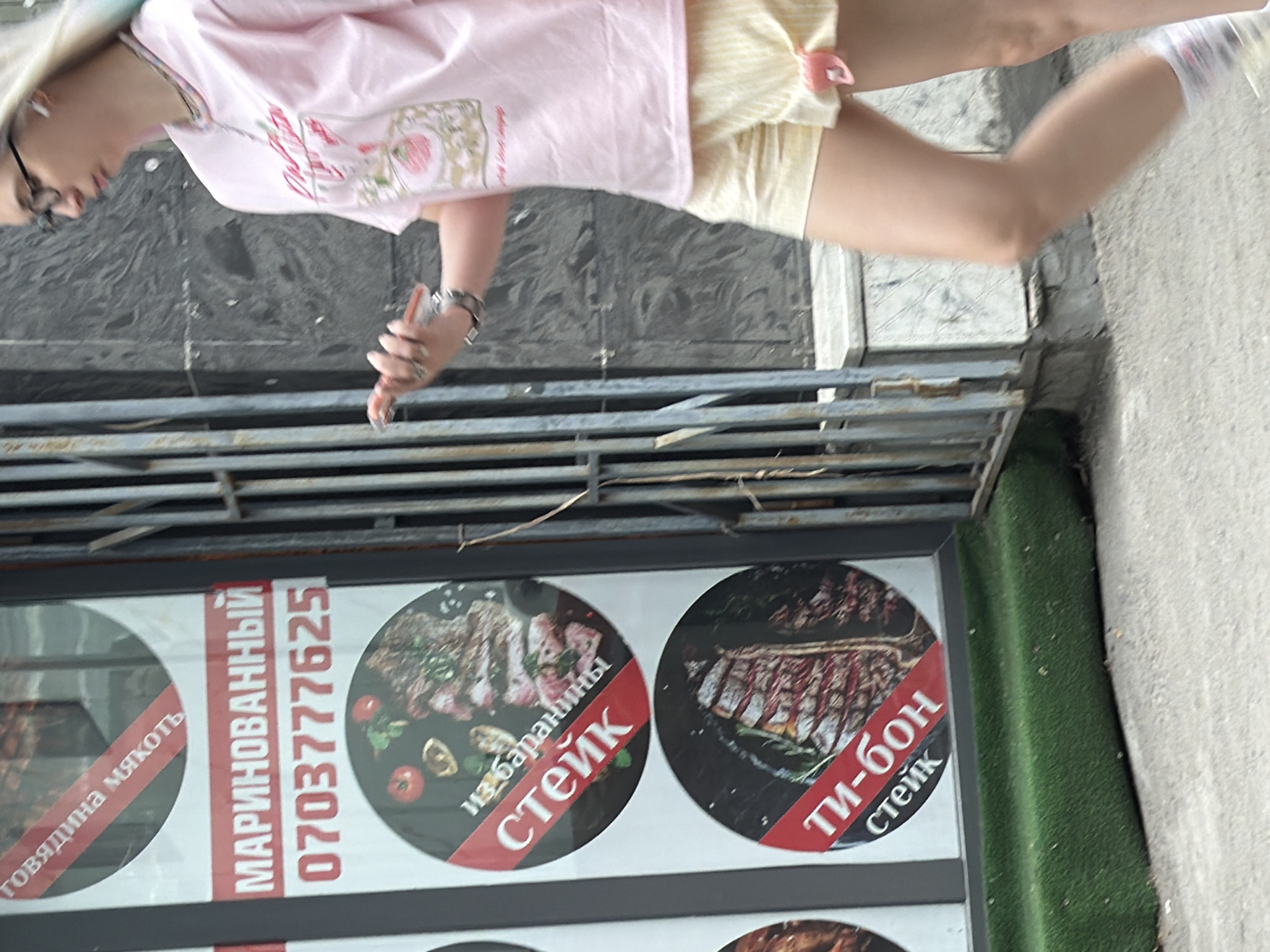 A young Caucasian woman, seen from the side, walks past a storefront on a sunny day. She has blonde hair with blue and green streaks and wears glasses, a pink t-shirt with a strawberry graphic, and yellow striped shorts. She is holding a cell phone in her right hand. The storefront displays signs with images of steak. The background consists of a metal gate and a marble-patterned wall. The ground is paved with concrete.