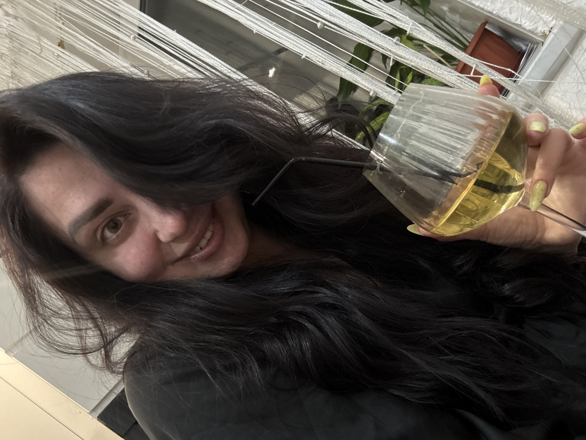 A woman with long, dark hair smiles at the camera while holding a glass of yellow liquid with a straw. The location appears to be indoors, possibly near a window with white string curtains and a potted plant. The city is unknown.