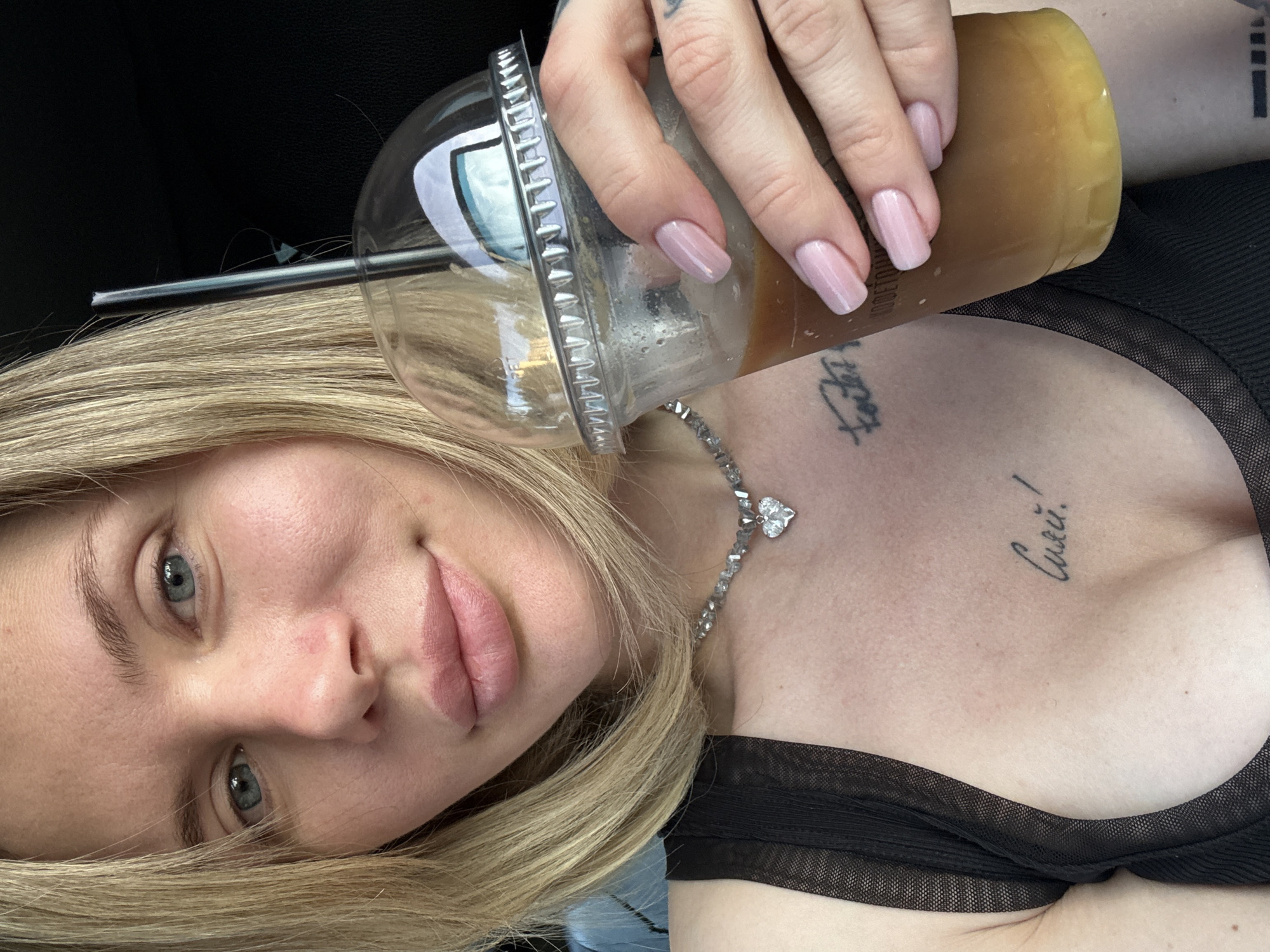 This close-up selfie shows a young woman looking directly at the camera, her head tilted. She has blonde hair, light blue eyes, and light pink lipstick. Her right hand, featuring light pink manicured nails, holds a clear plastic cup with a lid and straw, containing a light brown beverage, possibly iced coffee or tea.
She is wearing a black top with a mesh or sheer fabric texture. A delicate silver-colored necklace with a heart-shaped pendant is visible around her neck. Several tattoos are present: one on her upper left chest reads "Сшей!" (Russian for "Sew!" or "Stitch!"), and another partially visible on her inner right arm/bicep starts with "ro" or "ko" followed by indistinct letters.
The background is dark and out of focus, suggesting an indoor setting, possibly a car interior. The lighting is soft and natural. The image conveys a casual and personal moment. The provided location context for this media file is Gorno-Altaysk, Russia.