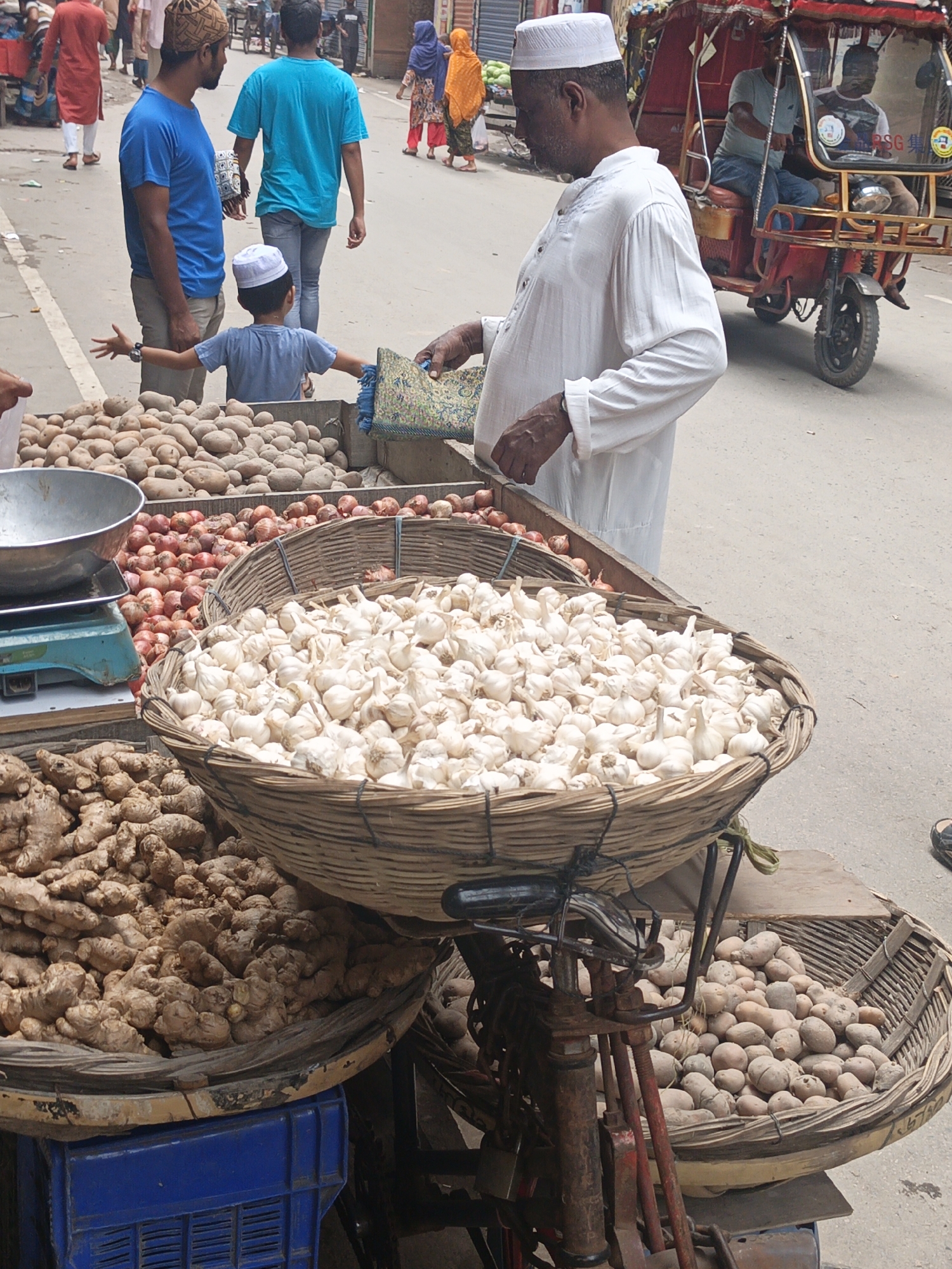 The image shows a street market scene in an outdoor setting, likely in India, given the presence of an auto-rickshaw. In the foreground, a variety of produce is displayed in large baskets. The most prominent is a woven basket overflowing with heads of garlic. To the left, a basket filled with ginger roots sits atop a blue plastic crate. Behind these, a larger basket contains a pile of small red onions, and further back, an even larger collection of potatoes is visible. A metal scale with a bowl is also present.
Several people are in the scene. A man in a white, long-sleeved tunic and a white cap is standing near the garlic, appearing to interact with the market stall. A young boy in a blue shirt and white cap is positioned in front of a pile of potatoes, with his arms outstretched. Two other men are seen in the background; one is wearing a blue t-shirt and the other a dark shirt. An auto-rickshaw, painted red, is in the middle ground, with at least two individuals inside, one appearing to be driving. Other figures and market stalls are faintly visible in the background, suggesting a bustling marketplace. The ground is paved with asphalt or concrete.