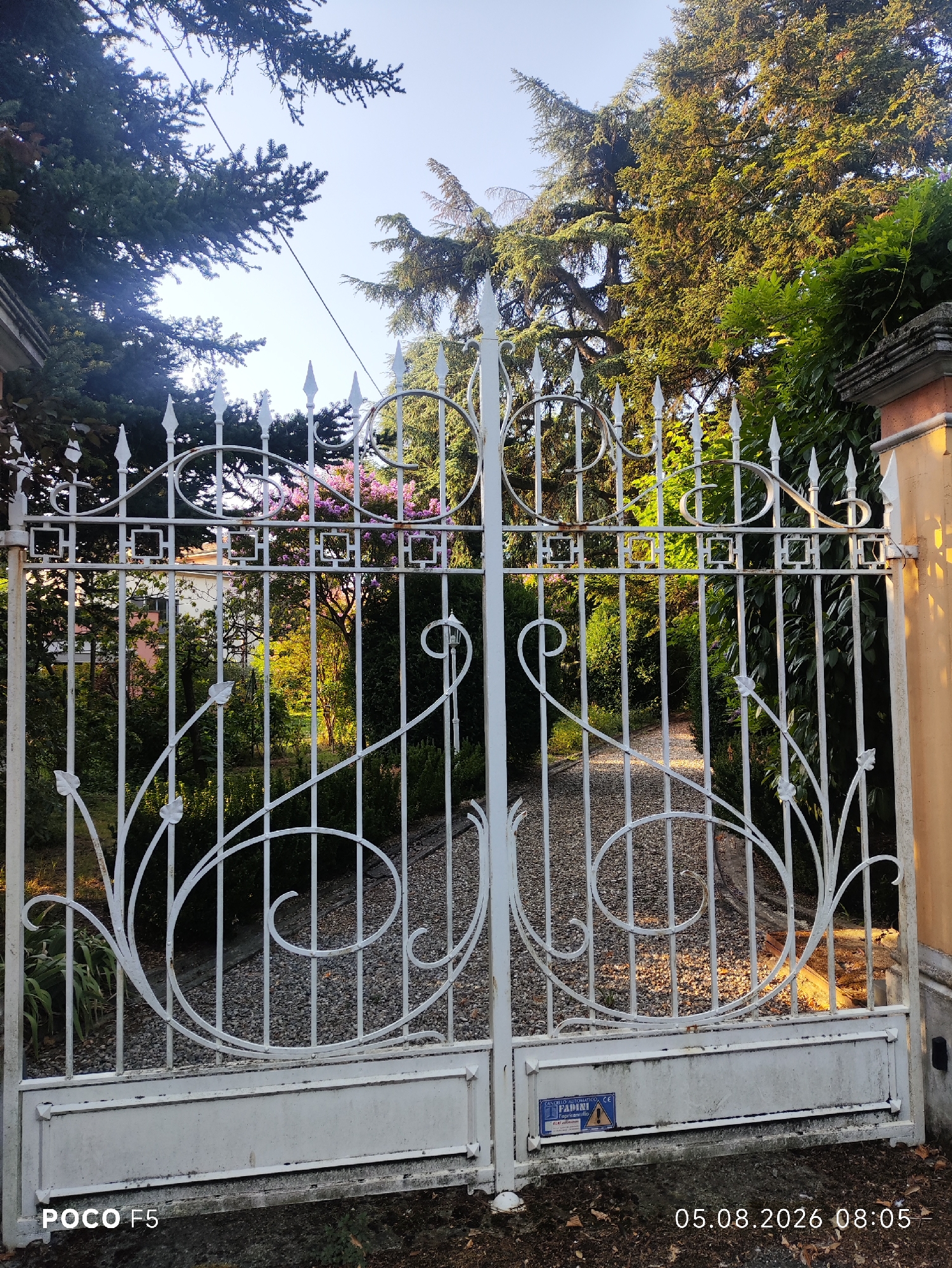 The image shows a white wrought iron gate with decorative scrollwork and pointed finials, partially open to reveal a gravel pathway leading into a garden. The gate is set in front of lush green foliage and trees, with hints of pink flowers visible in the background. On the lower section of the gate, a blue and yellow sticker with text that appears to be in Italian, possibly related to a gate automation system, is affixed. The timestamp "05.08.2026 08:05" and "POCO F5" are overlaid on the image. The overall scene suggests an entrance to a private residence or estate. Given the metadata and visual context, this is likely a residential property in Collecchio, Italy.