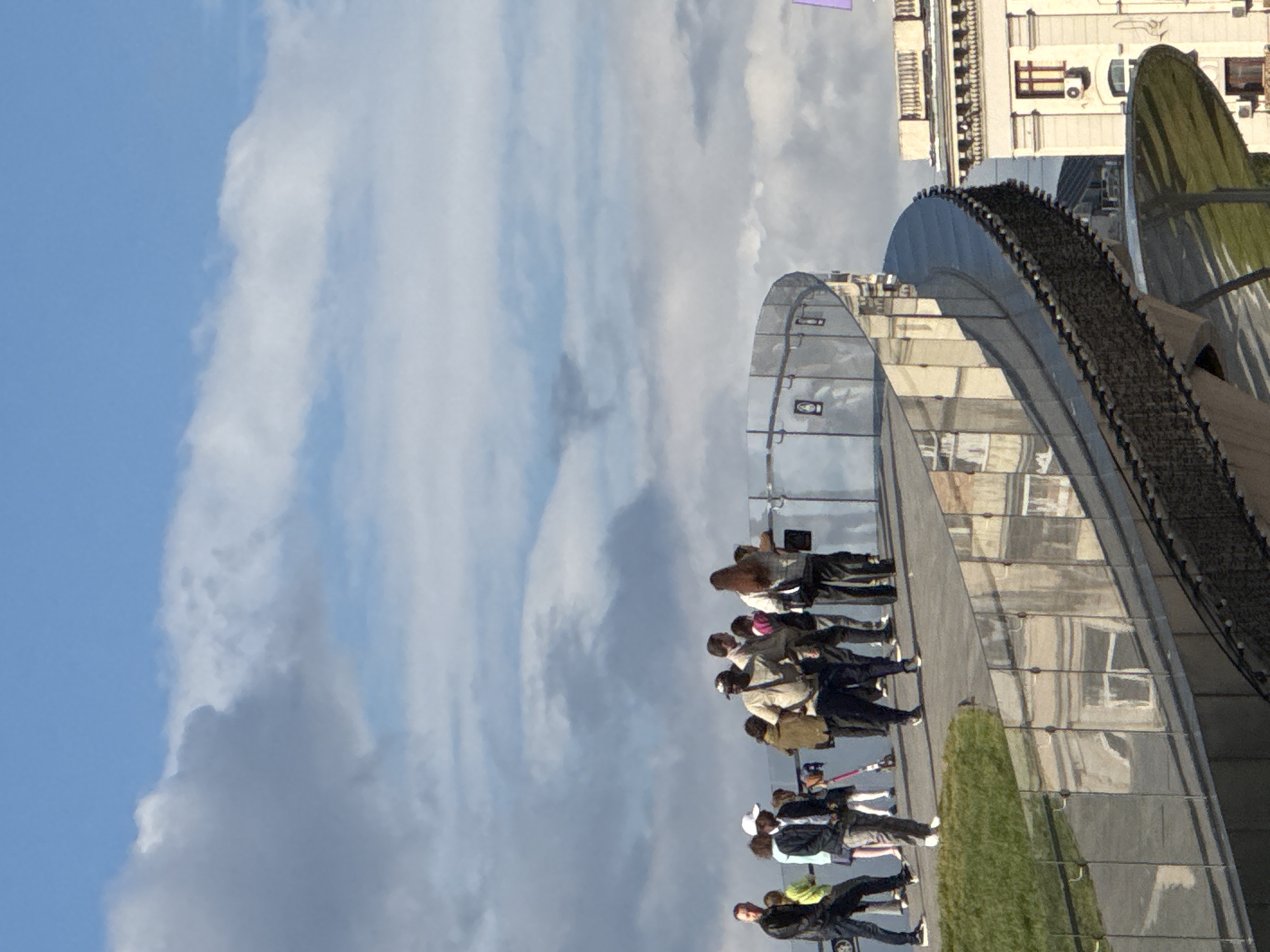 The image shows a group of people walking on a structure with an arch design, against a backdrop of a cloudy sky and buildings. The location is likely the Zaryadye Park in Moscow, Russia, with the "Floating Bridge" visible. The people are in various casual attire and seem to be enjoying the view. The structure they are on has a metallic framework and glass panels. The ground nearby is covered with grass.
