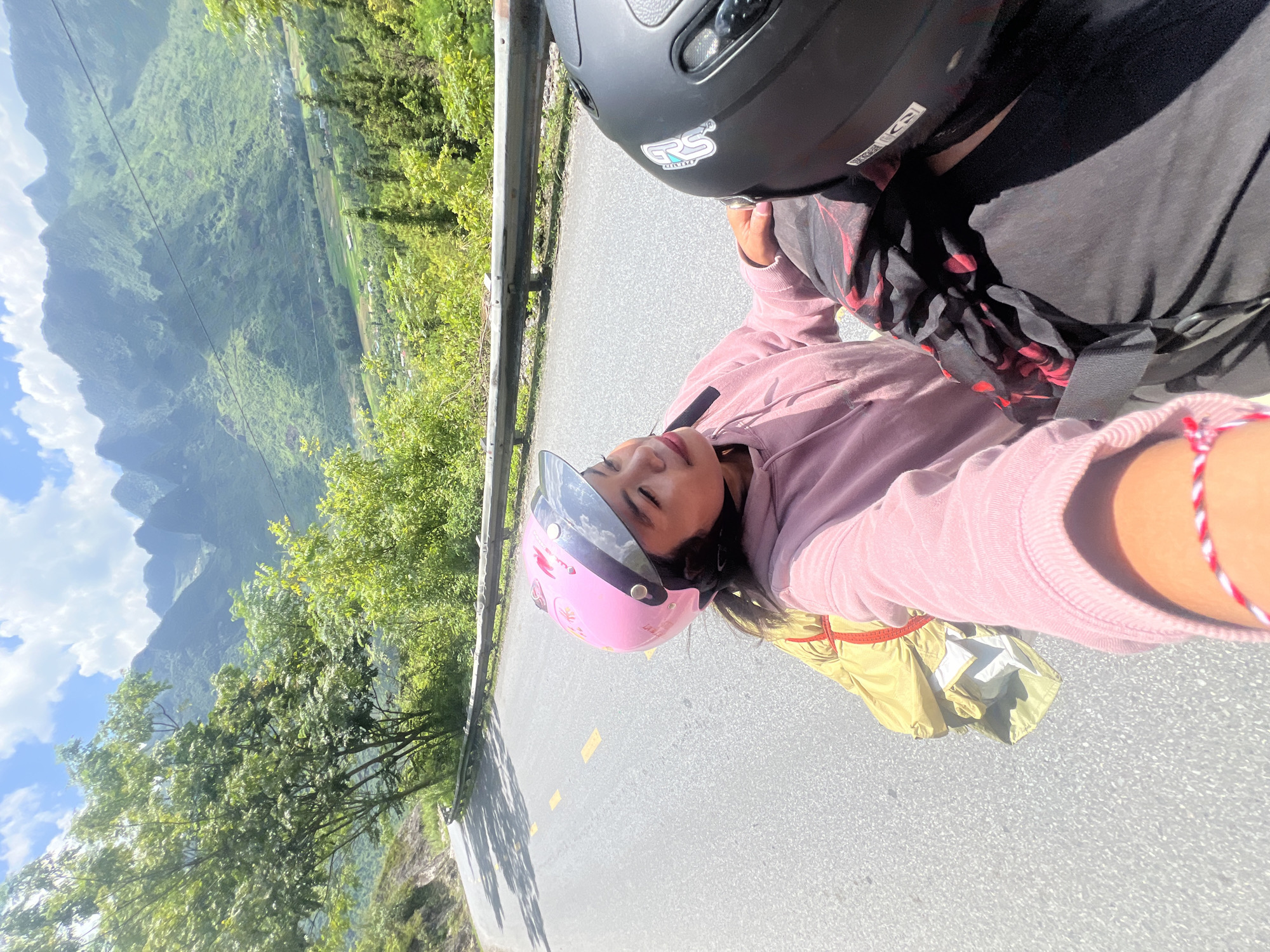 The media file captures a selfie taken by two individuals on a road, likely in Lùng Tám Commune, Vietnam, given the surrounding lush green mountains and valleys. The scene is set outdoors during the daytime, under clear blue skies with some clouds. The road appears to be paved and is bordered by a metal guardrail and dense vegetation.
The primary subjects are two people on what seems to be a motorbike. The person in the foreground, wearing a pink hoodie and a pink helmet, is looking towards the camera, with their eyes closed and a serene expression. They are also wearing a yellow jacket underneath their hoodie, and a red and white bracelet is visible on their wrist. Behind them, the head of another person wearing a black helmet is visible.
The activity appears to be a motorbike ride through a scenic mountainous region. The backdrop showcases the dramatic landscape with steep, verdant mountains, some of which are covered in trees and possibly terraced fields. The bright sunlight suggests it's a clear and pleasant day, ideal for travel. The overall mood conveyed is one of relaxation and enjoyment of the journey and the breathtaking scenery.
There is some visible text on the helmets. The black helmet has a sticker with "GVS" and some other lettering. The pink helmet has several stickers with cartoon-like images. The road has yellow painted lines.