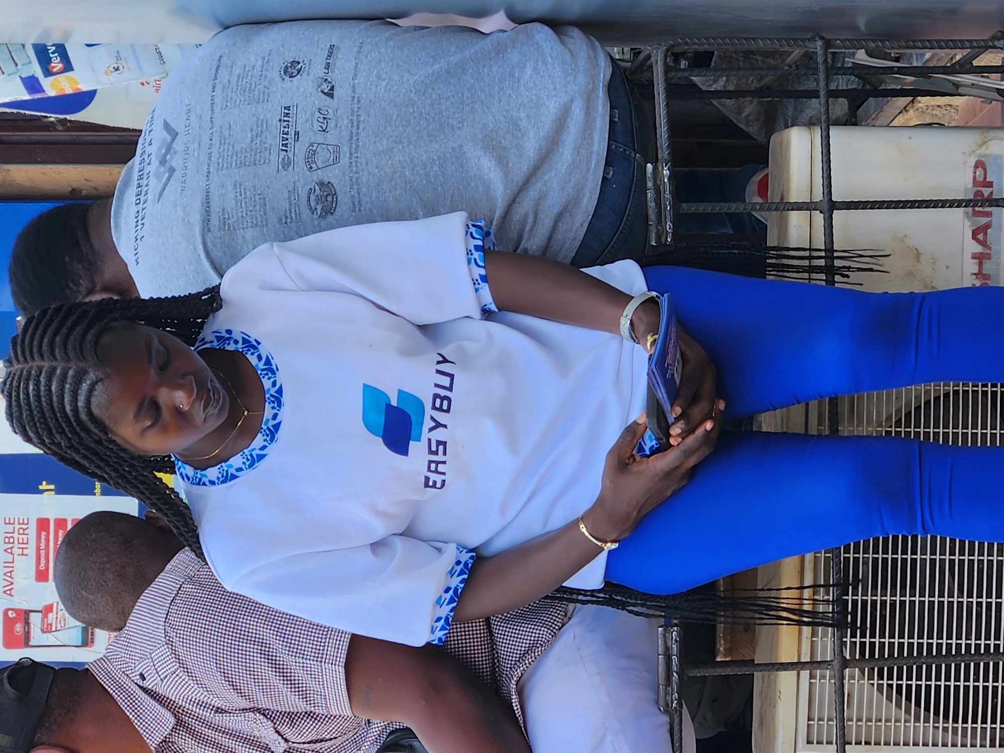 The image shows a woman wearing a white t-shirt with the "EASYBUY" logo and blue leggings, sitting on what appears to be a metal structure. She is holding a mobile phone and looking at it. Behind her is another person with their back towards the camera, wearing a gray t-shirt with multiple logos and text on it. To the left, another individual is partially visible, wearing a checkered shirt and a black cap. In the background, there is a sign mentioning "AVAILABLE HERE" for "Deposit Money." There are also other signs and materials in the background, including mentions of "Verve" and "airt." In the upper right corner, part of a "SHARP" branded appliance is visible. The location is likely Jos, Nigeria, based on the "EASYBUY" logo, which is associated with financial services in Nigeria.