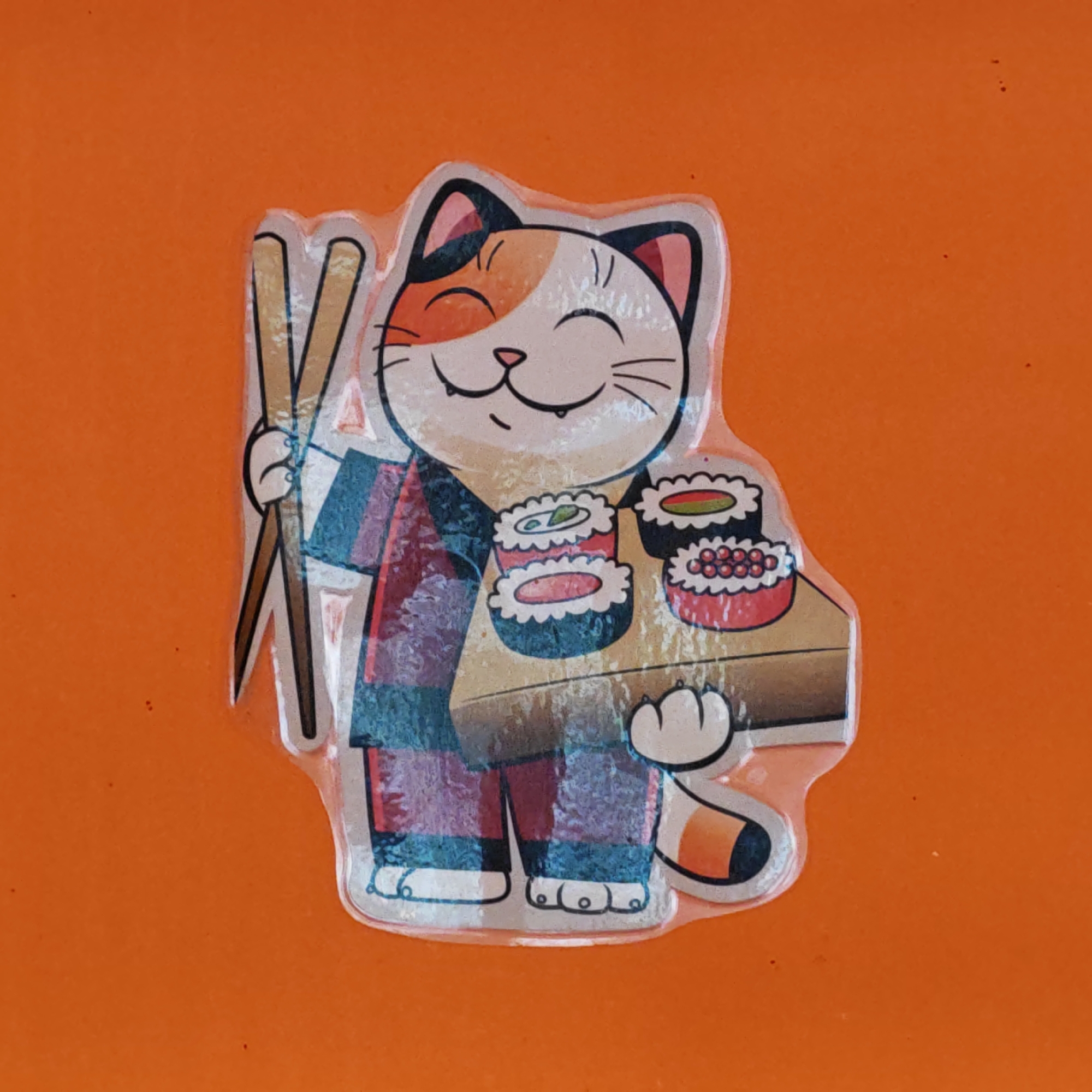 A sticker of a cartoon cat waiter is centered on an orange surface. The cat has a calico pattern on its head and wears a traditional Japanese garment, possibly a kimono or yukata, in shades of red and blue. The cat holds chopsticks in one hand and a tray of sushi in the other. There is no indication of the location being Rostov, Russia.
