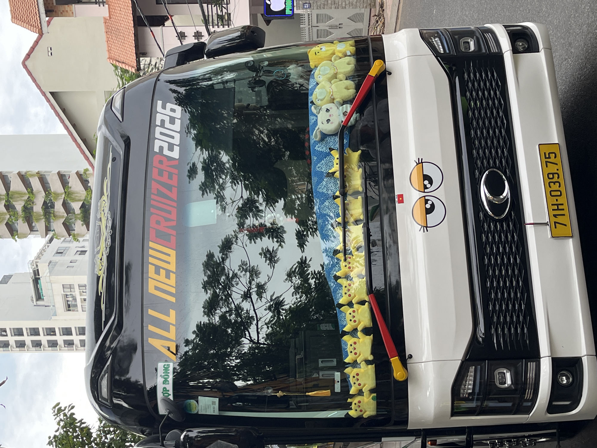 A white and black bus is parked on a street in Nha Trang, Vietnam. The bus has a large windshield adorned with several yellow stuffed Pikachu toys and other plush animals. A blue patterned fabric is visible behind the plush toys. A license plate that reads "71H-039.75" is affixed to the front of the bus. Above the windshield, the words "ALL NEWCRUIZER 2026" are prominently displayed in colorful lettering. Buildings, including a tall white building with green vines, are visible in the background. The sky is overcast.