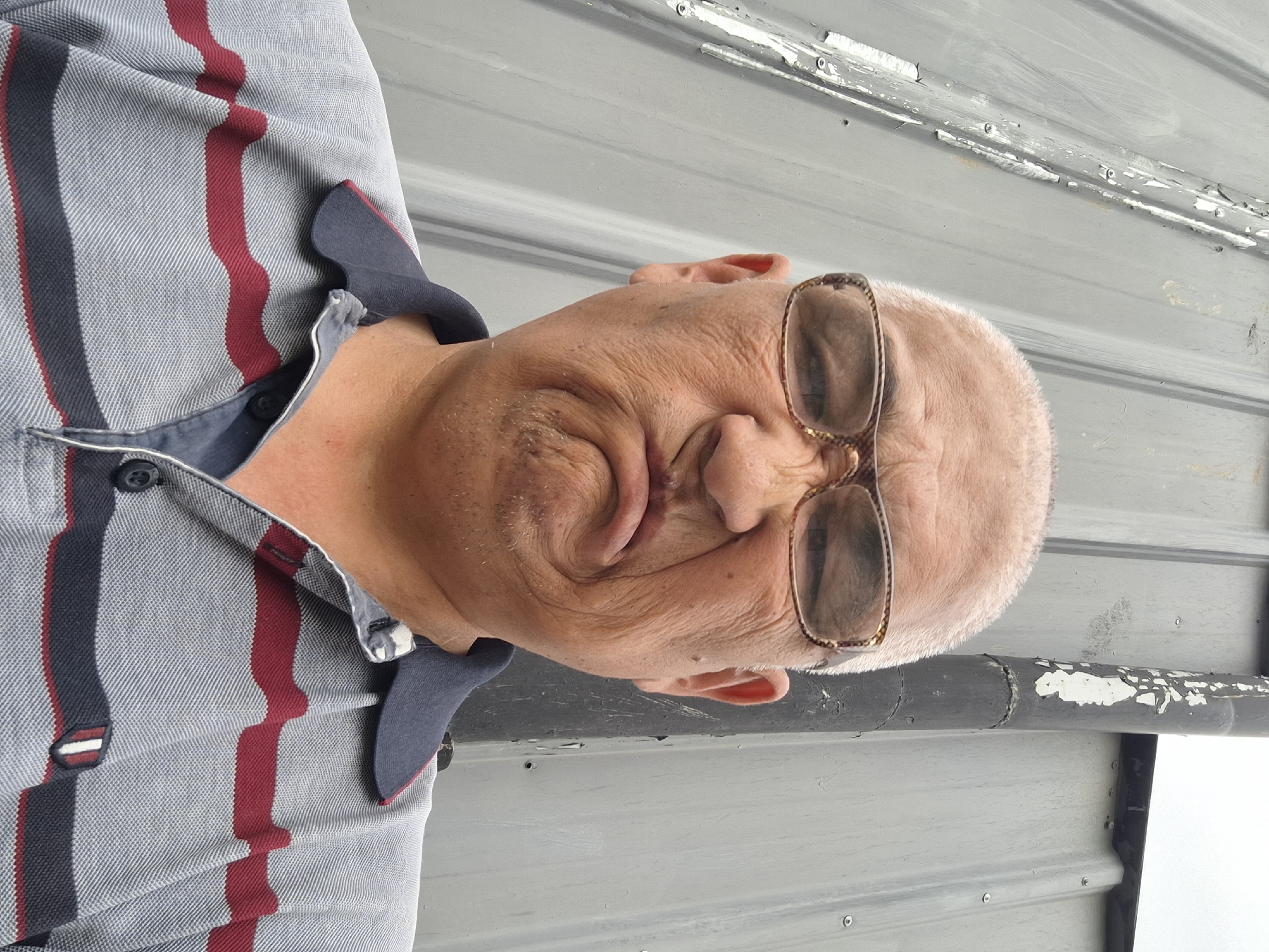 The image is a close-up, rotated portrait of an older man wearing glasses and a collared shirt with red and dark blue stripes. The man's head is tilted, and his face is partially obscured by his glasses. His skin appears wrinkled, and he has a light stubble. He is looking downwards. The background consists of a grey, corrugated metal surface, possibly a wall or a roof, with peeling paint in some areas. There is no discernible activity or event taking place. The lighting suggests it is daytime, and the background implies an outdoor or industrial setting. No text is visible in the image.