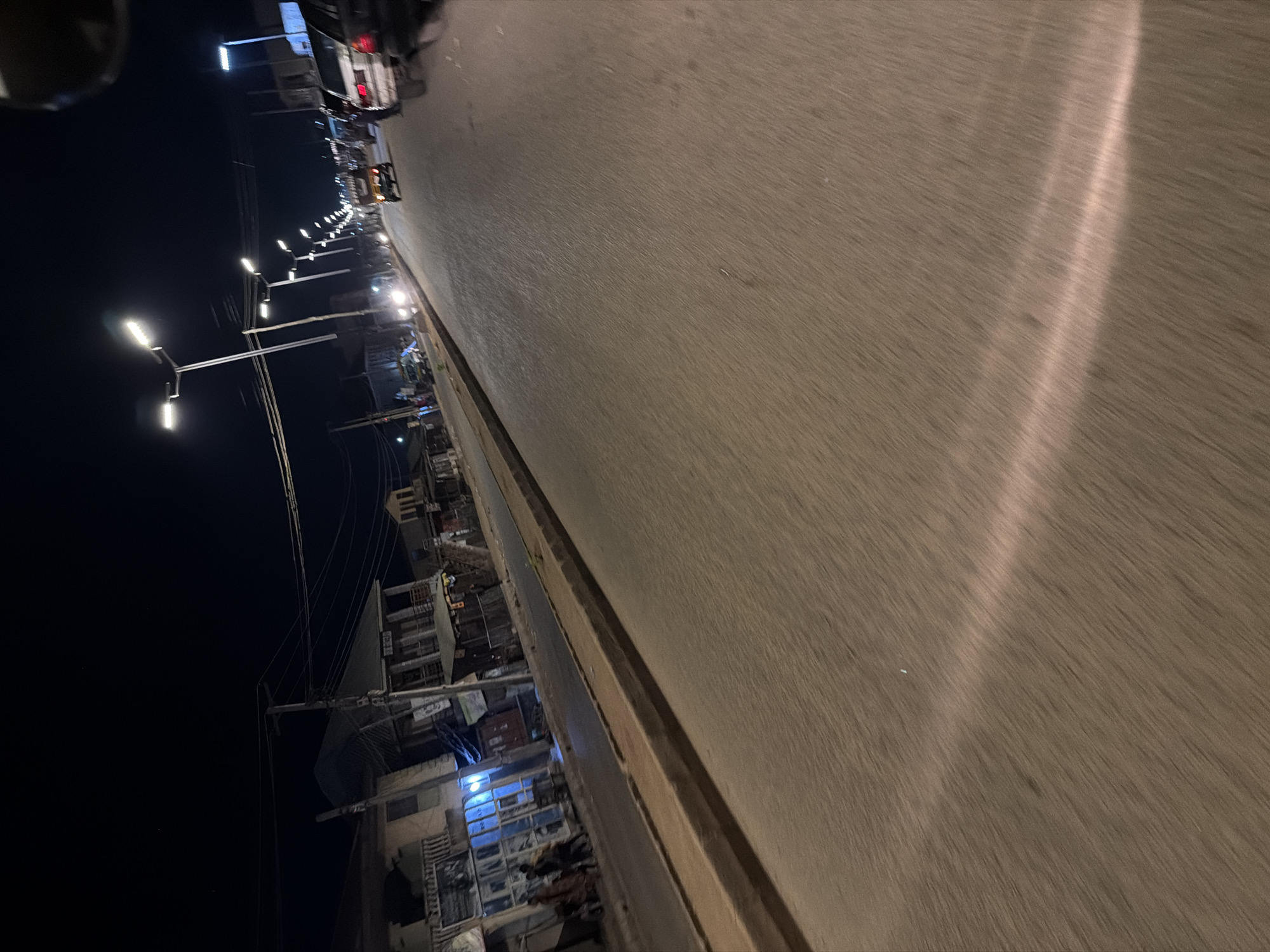 The image depicts a street scene at night in Bukuru, Nigeria. The street is paved and illuminated by streetlights. On the left side of the street, there are buildings with lights visible in the windows. Several people are walking on the sidewalk in front of these buildings. Further down the street, vehicles are visible, including cars and what appears to be a yellow van. The road surface shows motion blur, indicating that the photograph was likely taken from a moving vehicle. The overall atmosphere is that of an active urban environment at night.