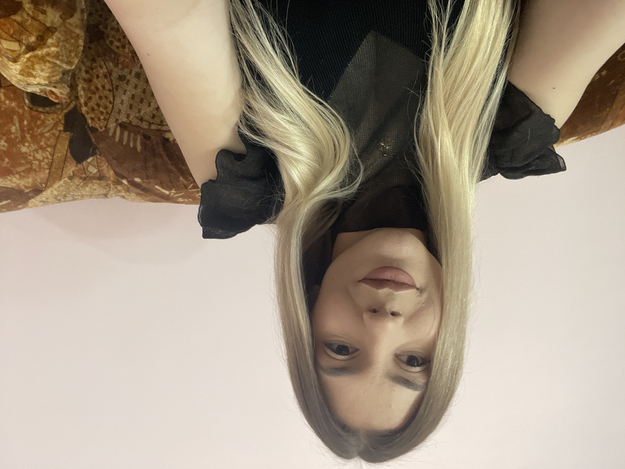 A medium-shot, indoor photograph shows a woman from the chest up, with the image rotated 180 degrees. The woman has fair skin, light brown eyes, and light pink lips. She is wearing a black top that appears to have some sheer elements. Her hair is long, straight, and blonde.
Behind her and to her right is a couch or other furniture with a brown patterned fabric. The background is a plain, light pink or off-white color.
There is no information to suggest the location is in Burgas, Bulgaria. The image is devoid of any landmarks or identifying characteristics that would lead to this conclusion.