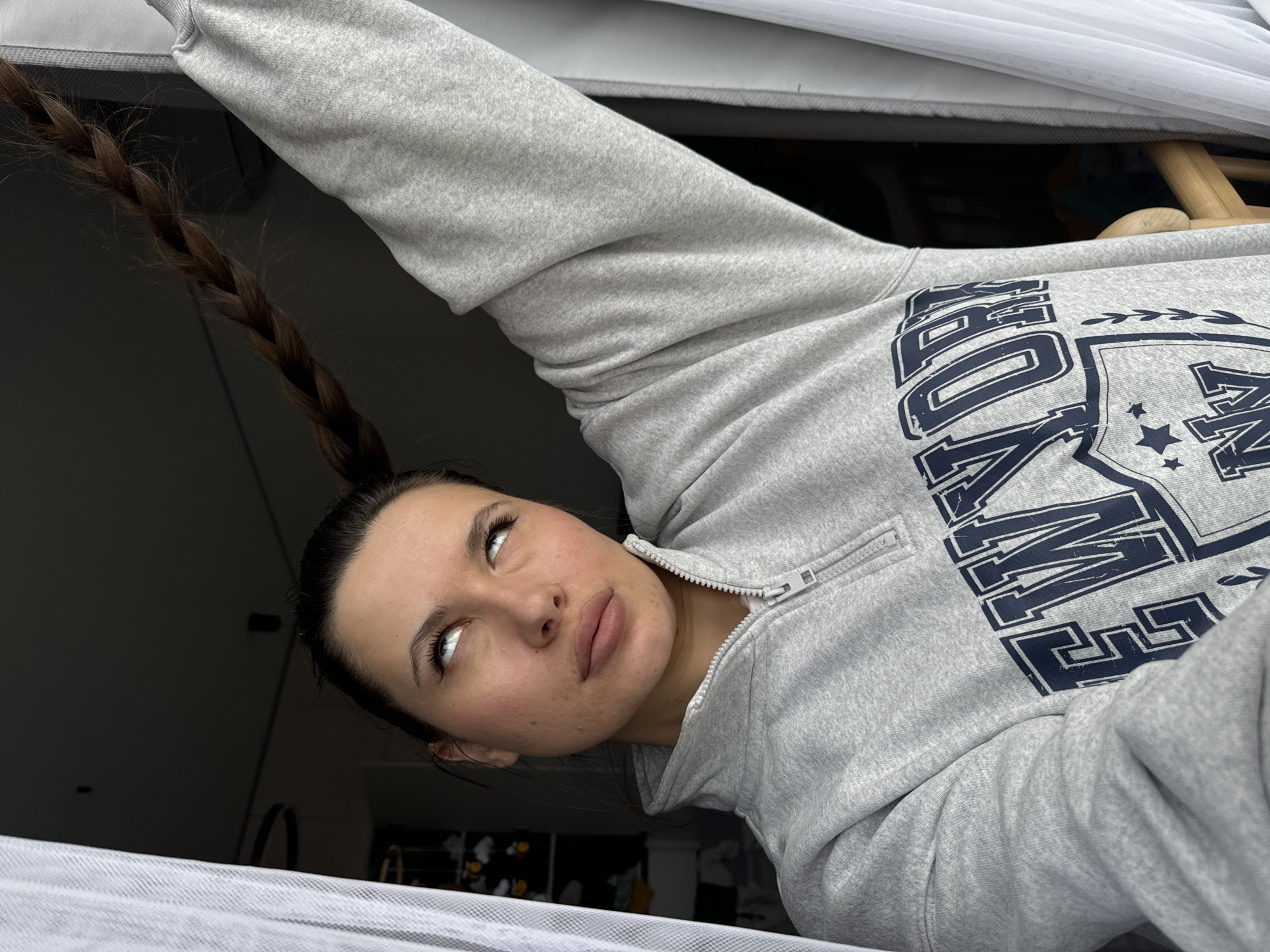 A young woman with fair skin and a long brown braid stands inside a room, looking upwards. She wears a gray zip-up sweatshirt with dark blue lettering and a shield design on the front. The background includes dark, out-of-focus areas, suggesting the interior of a room. A white sheer fabric is visible in the foreground. The photograph appears to be taken from a low angle, looking up at the subject. The image does not provide sufficient information to determine the exact location in Odintsovo, Russia.