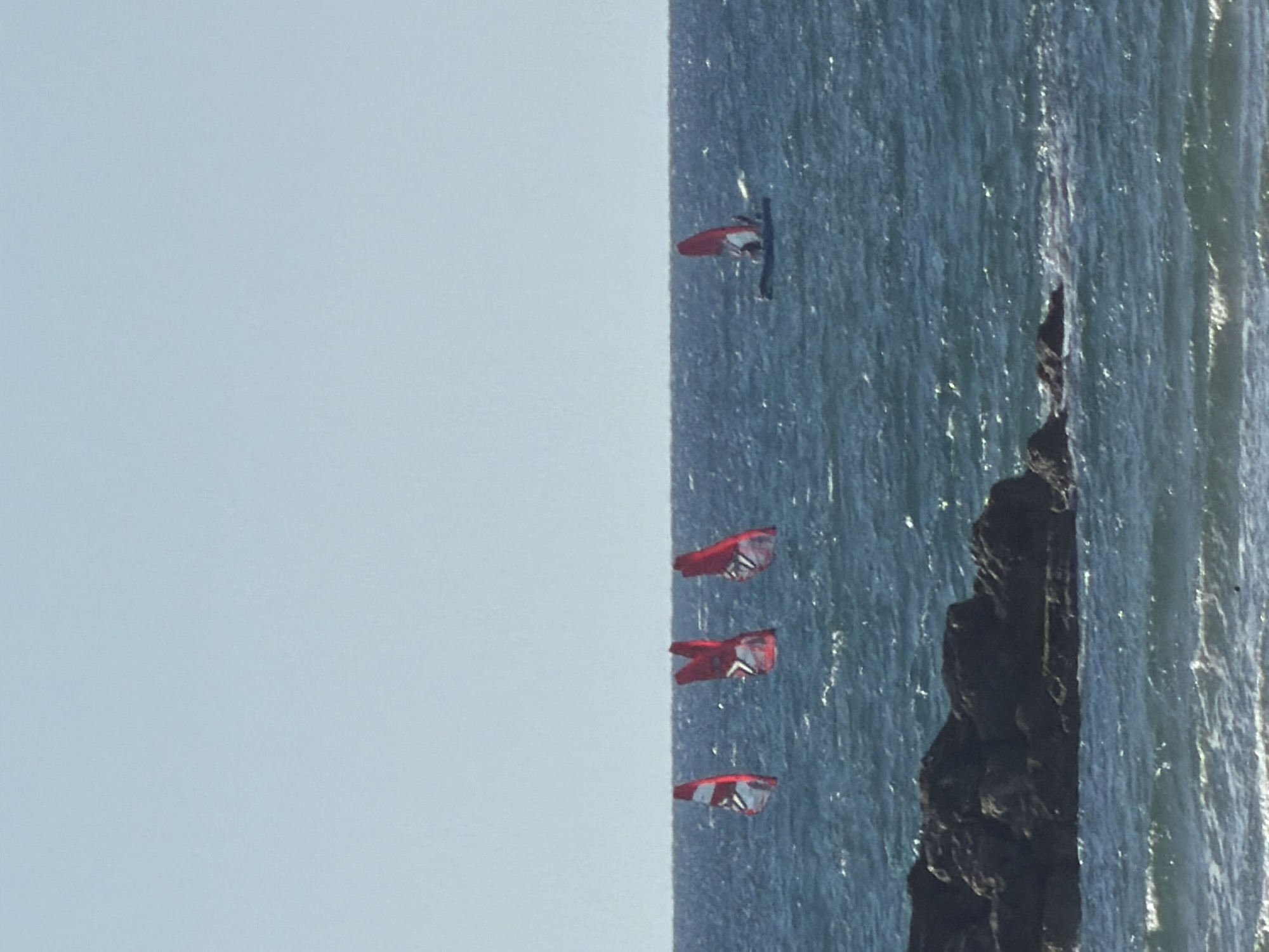 The image shows a vertical shot of the sea adjacent to a rocky outcrop. Four red and white kites are visible in the water. A person is standing in the sea near the top kite. The kites and person are positioned along the edge where the sea meets a pale blue sky. Based on the kitesurfing activity and coastal features, this scene could be located near the coast of Tel-Aviv, Israel.