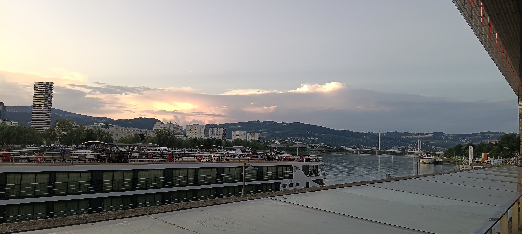 In Linz, Austria, a wide shot captures a twilight scene by a river. In the foreground, the upper deck of a cruise ship is visible, with tables, chairs, umbrellas, and people seated and standing. The ship has multiple levels of windows. To the left, a modern high-rise building stands against a backdrop of rolling hills and other urban structures. Across the river, more buildings and a suspension bridge are visible in the distance, with hills forming the horizon. The sky is filled with dramatic clouds illuminated by the setting sun, casting warm hues. The right side of the image shows the edge of a contemporary building with illuminated vertical strips.