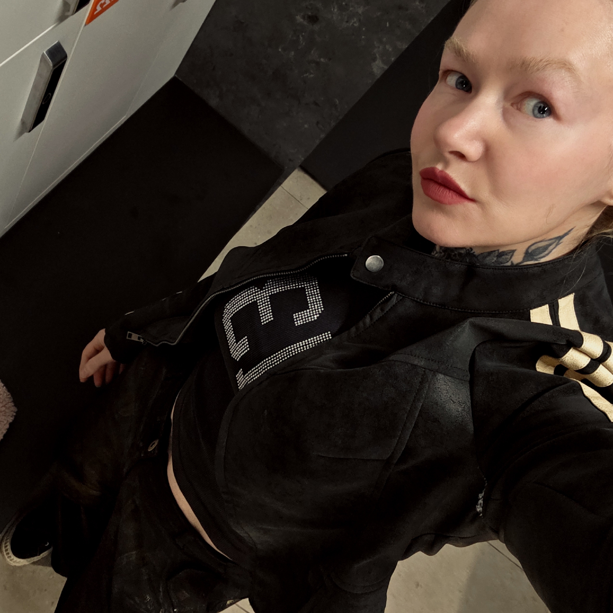 The image is a high-angle, downward-facing selfie featuring a fair-skinned individual, likely female, with blonde hair and eyebrows, and striking blue eyes. She looks directly at the camera with a neutral expression, enhanced by red lipstick. A black ink tattoo depicting foliage is visible on her neck, just below her ear.
She is dressed in a dark outfit, including a black jacket, possibly suede or leather-like, with a zipper and a silver snap button at the collar. The jacket has gold or light-colored striped accents on the sleeves or shoulders. Beneath it, she wears a black top featuring a large, stylized letter or symbol (resembling an "E" or "F") intricately formed by small silver or white studs across the chest. A strip of her midriff is visible. Her left hand is visible by her side, and a black sneaker with a white sole can be seen on her left foot.
The setting is an indoor, modern space. The background consists of clean white walls, one featuring a metallic handle or fixture and a partially visible orange triangular symbol. The floor is predominantly dark, possibly a dark carpet or polished surface, with a lighter-colored floor section visible at the bottom. A fluffy, light-colored object is partially visible on the far left. The overall environment suggests a contemporary building, possibly a commercial or public area.
