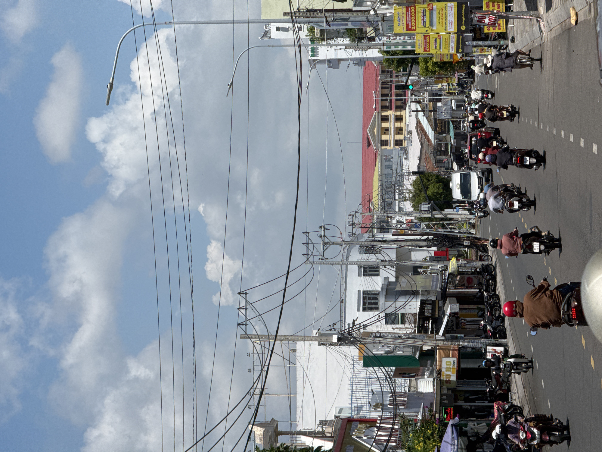 The image shows a street scene in Phú Thủy, Vietnam, on a bright, cloudy day. The street is busy with traffic, predominantly motorcycles. Several people are riding motorcycles, some with helmets. Buildings line both sides of the street, featuring a mix of commercial establishments with signage and residential structures. Overhead, a complex network of electrical wires crisscrosses the sky. The road has yellow lane markings. A white van and a few cars are also visible in the traffic. The buildings have various architectural styles and colors, including white facades, red roofs, and some with yellow accents. Billboards and streetlights are also present. The overall impression is of a bustling urban environment in Southeast Asia.