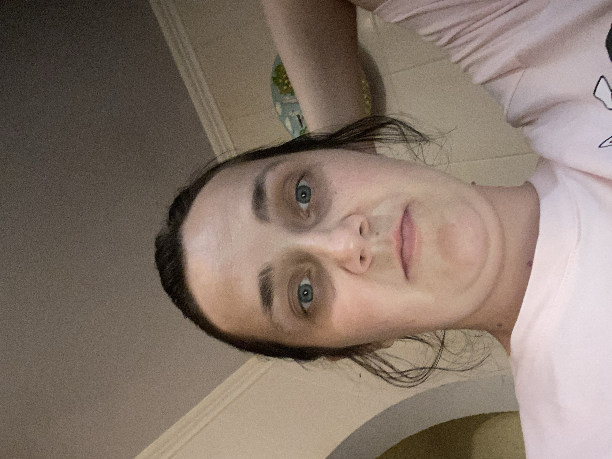 A low-angle, close-up shot shows a person from the shoulders up, facing the camera. The individual has dark hair pulled back, with some strands loose around the face. Their eyes are blue, and there are dark circles beneath them. The person's mouth is closed, and their chin is visible, with a slight double chin. They are wearing a pink t-shirt. The background consists of a beige wall and ceiling, with a lighter beige trim along the top. A curved archway is visible on the left side of the frame. Part of a person's arm and leg are also visible on the right, as well as a decorative plate on the wall. The lighting appears to be artificial and from above. The location is unknown.