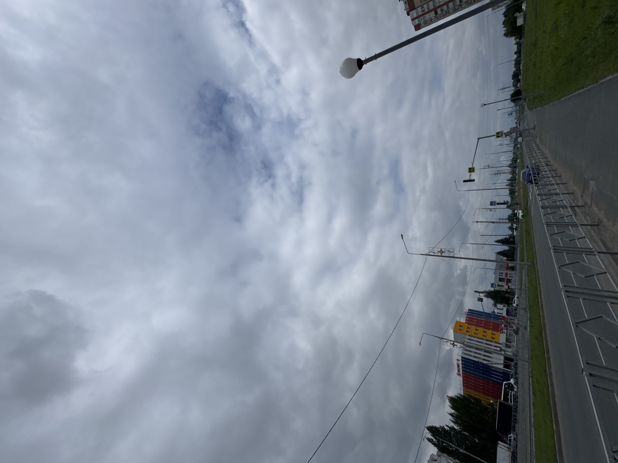 The image captures a view of a street in Tolyatti, Russia, under an overcast sky. A multi-lane road runs horizontally across the foreground, bordered by a sidewalk and a metal fence. On the left side of the road, a building with a distinctive facade featuring red, blue, yellow, and white panels is visible, identified by a sign that reads "МАГНИТ" (Magnit). Several cars and a truck are on the road. In the background, there are more buildings, streetlights, and trees. The sky is filled with grey, textured clouds, with some patches of lighter blue. The overall impression is of a typical urban environment on a cloudy day.