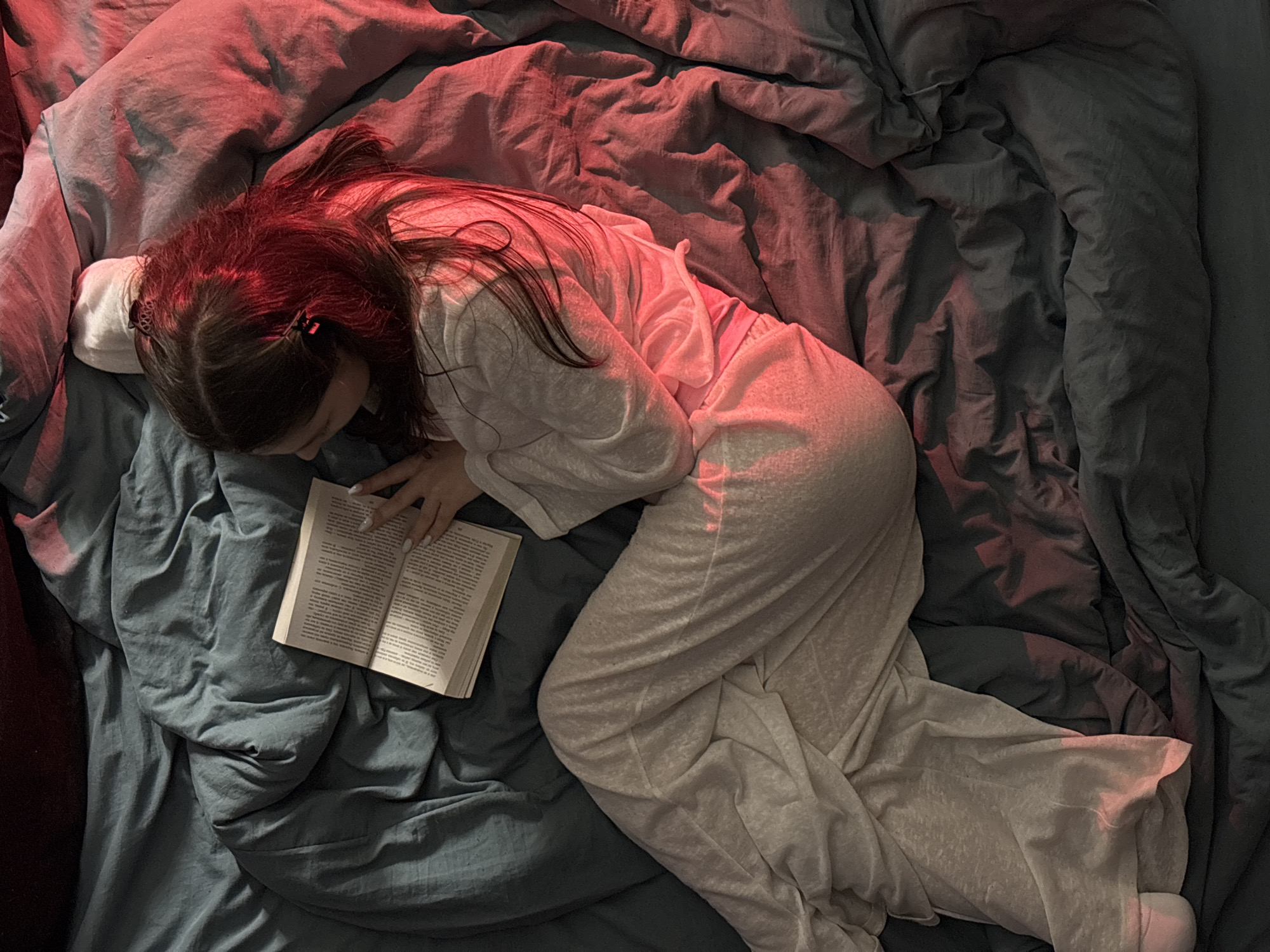 A young woman with long dark hair and two small black hair clips lies prone on a bed covered in a crumpled blue-grey duvet. She is wearing a long-sleeved white pajama set and reading an open book with both hands. The room is dimly lit with a red light source primarily illuminating the upper part of the frame. Location: Chaikovsky, Russia.