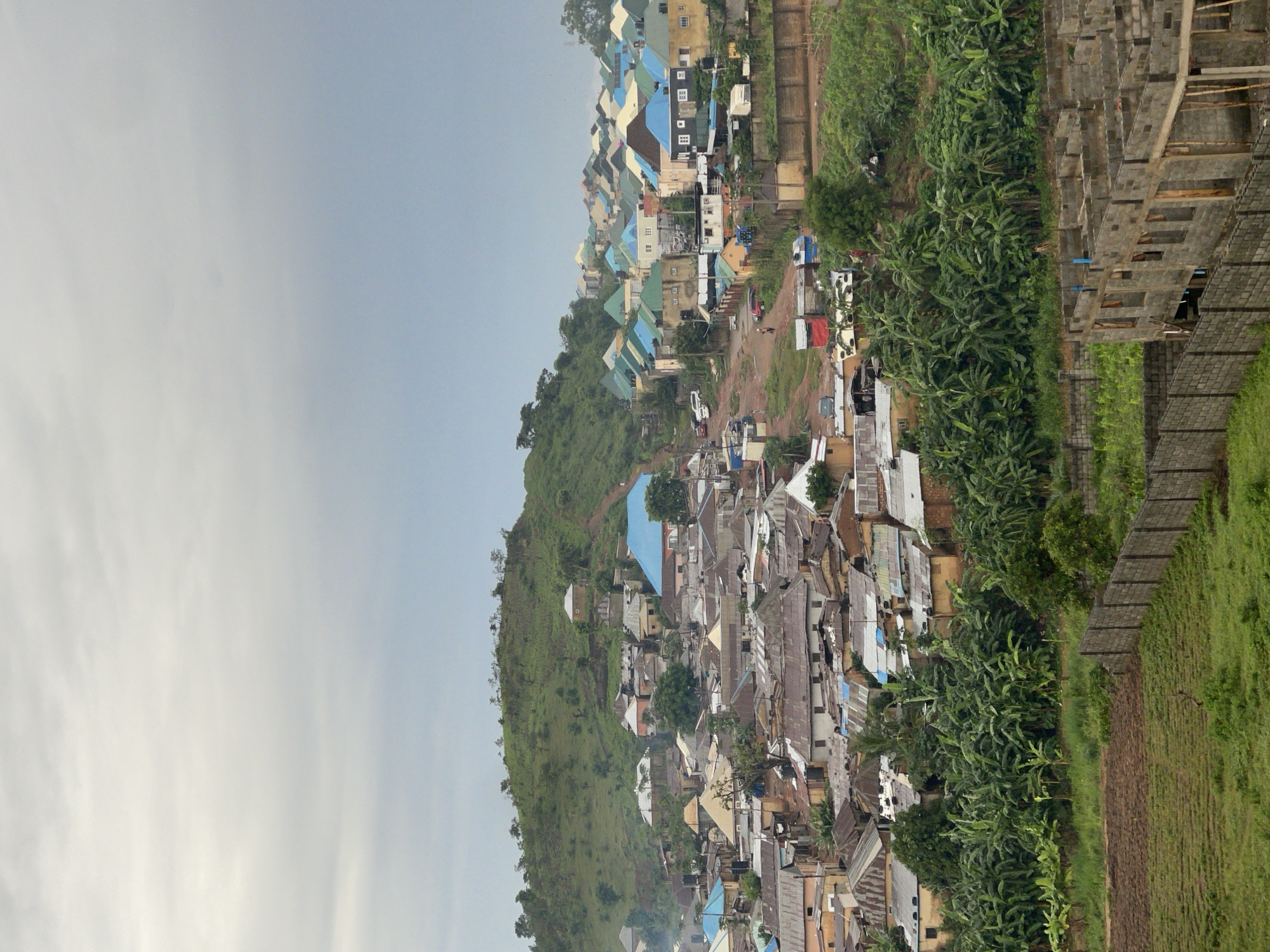 This is a photograph of a residential area in Abuja, Nigeria, showing a hillside covered in buildings and vegetation under an overcast sky. The structures are a mix of dwellings with corrugated metal roofs and some with colorful metal roofs in shades of blue and green. Many of the houses are situated on uneven terrain, giving the impression of a densely populated hillside community. Lush green trees, including what appear to be banana trees, are interspersed among the buildings and cover the lower slopes of the hill. In the foreground, a partially constructed building with exposed concrete framework is visible, enclosed by a brick wall. A dirt road winds through the settlement, with a few vehicles and individuals present, indicating ongoing activity. The overall scene depicts a vibrant, lived-in urban environment.