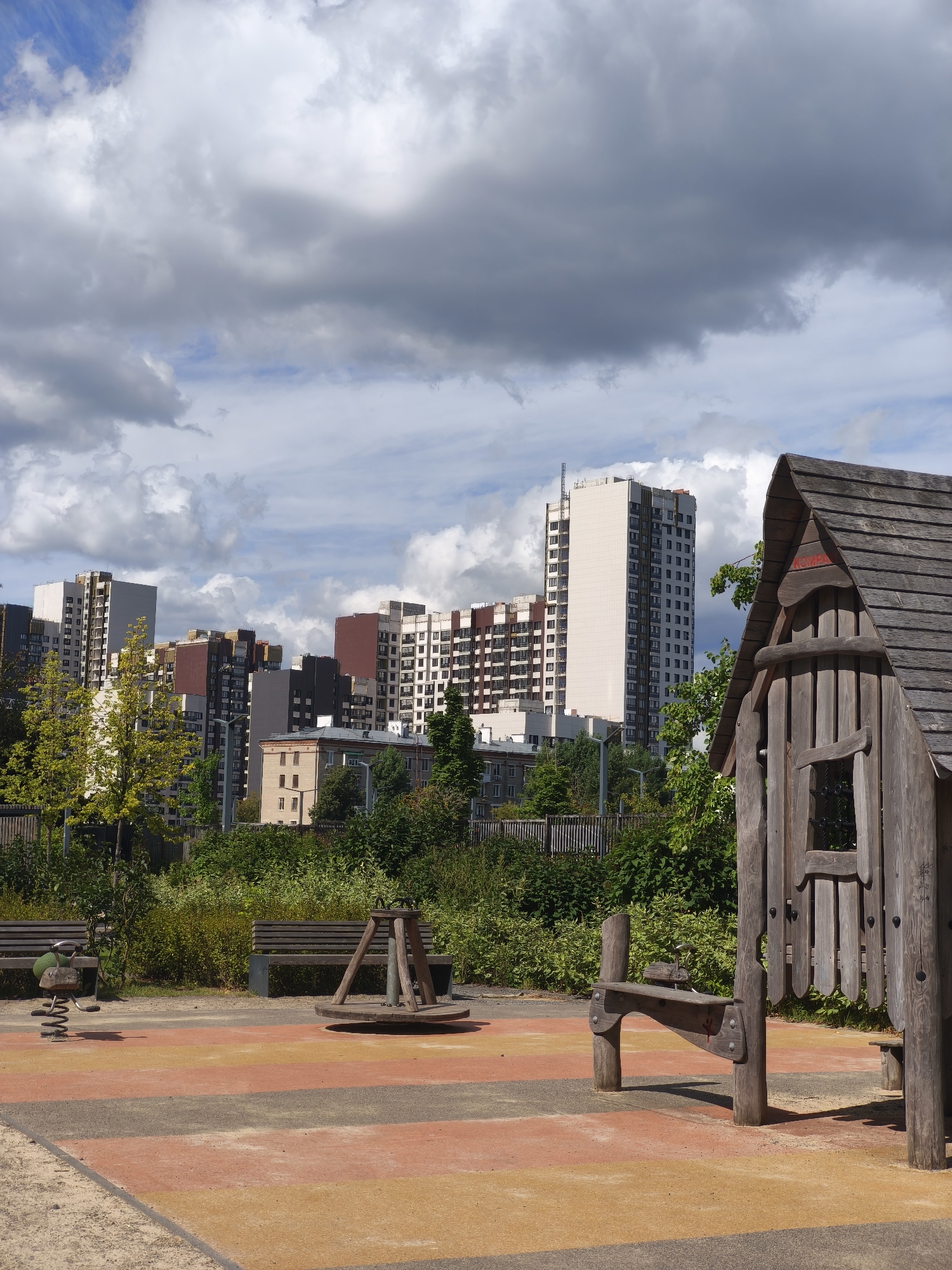 The image captures a public playground in Moscow, Russia, with a backdrop of contemporary high-rise apartment buildings. In the foreground, a wooden playhouse stands prominently, its rustic design contrasting with the modern architecture behind it. The playground itself features a colorful rubberized surface with striped patterns, a spring rider, a merry-go-round, and park benches. Lush green trees and foliage surround the play area, creating a natural buffer between the playground and the urban landscape. The sky above is filled with dramatic, grey clouds, suggesting a dynamic weather system. The overall scene depicts a blend of recreational urban space and residential development.