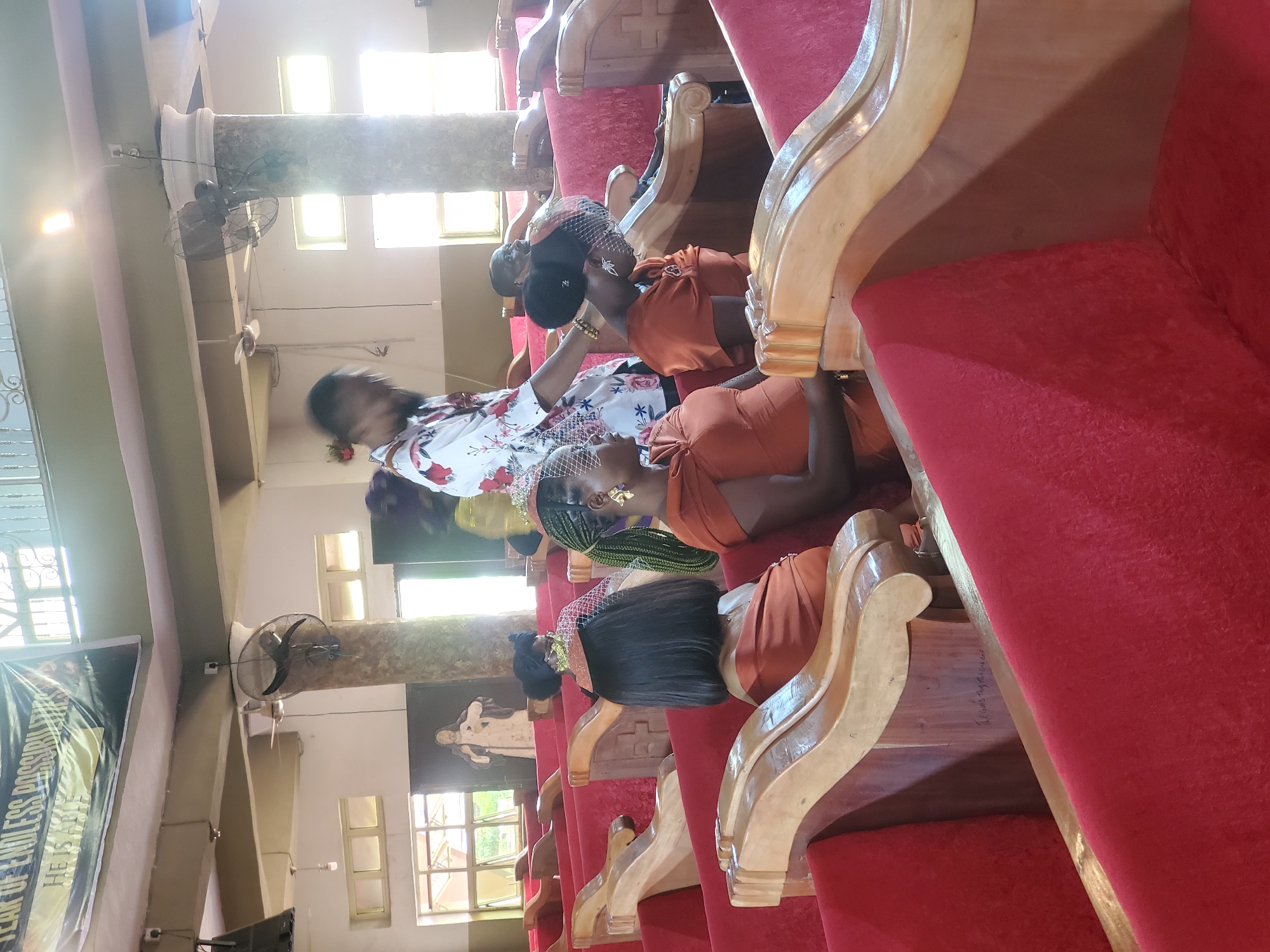 The image shows several women seated in rows of pews inside a building. The pews are red with light wooden frames. The women are wearing various outfits, including orange dresses and a white dress with floral patterns. Some are wearing head coverings with veils. The setting appears to be a church. Given the location in Onitsha, Nigeria, it is likely a church or religious institution in that city. The light is bright, filtering in through windows in the background. There are fans on the ceiling of the church.