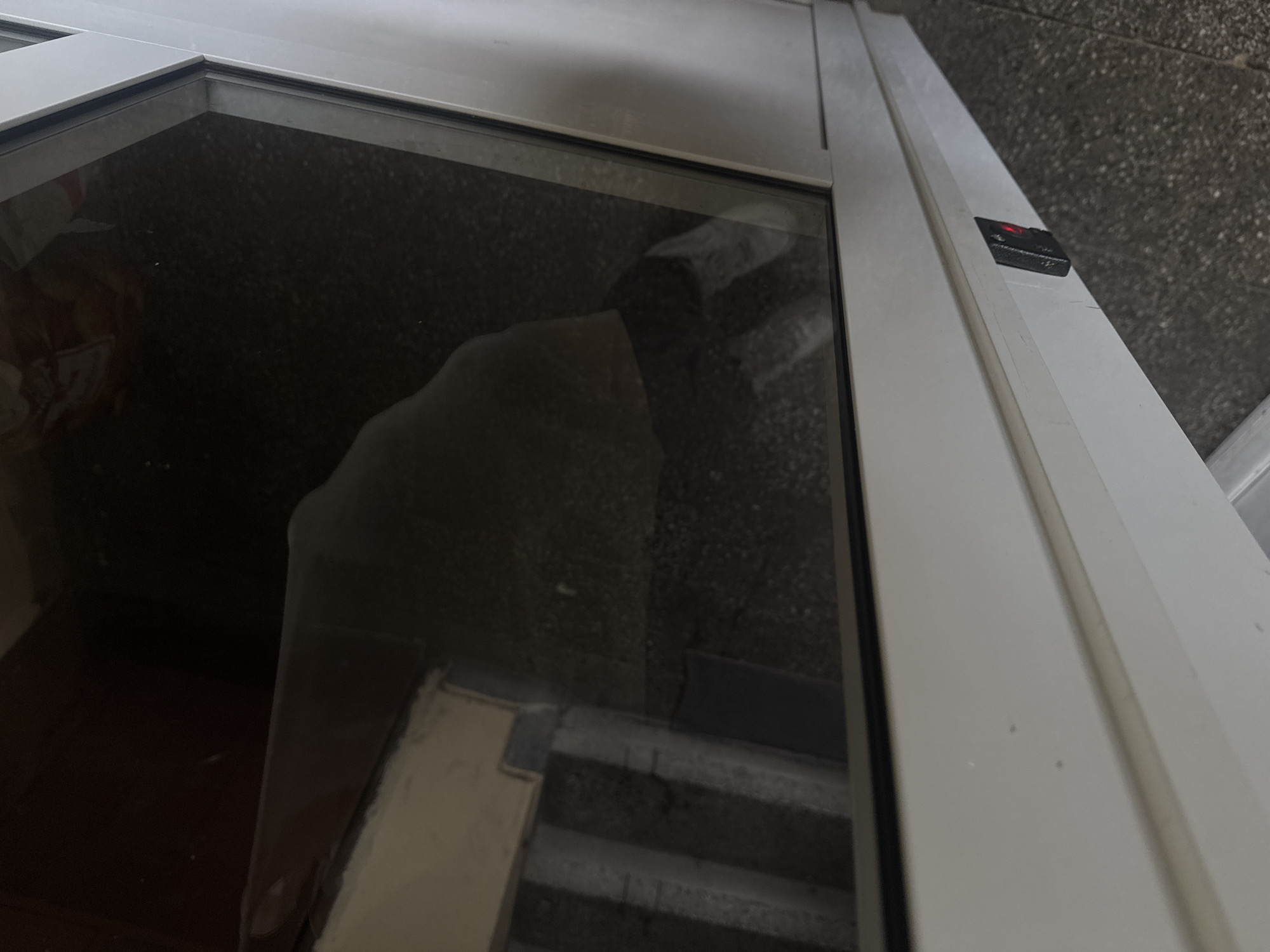 The image depicts a close-up, downward-angled view of a white, modern door, likely PVC or metal-framed, with a prominent glass panel. On the left vertical frame of the door, a black electronic access control reader is mounted, displaying a red illuminated indicator light. The immediate floor in the foreground, and visible through the bottom left of the glass, is a dark, speckled terrazzo or aggregate surface. Through the upper section of the glass panel, a section of a concrete or grey-painted staircase is visible, ascending away from the viewer. Reflections on the glass obscure a portion of the view, showing indistinct dark shapes and lighter smears, possibly internal architectural elements or reflections from the observation point. No individuals, actions, or interactions are observable. The environment appears to be an interior building space, such as an entrance or a stairwell landing. The image does not contain identifiable features to confirm a specific location within Varna, Bulgaria.
