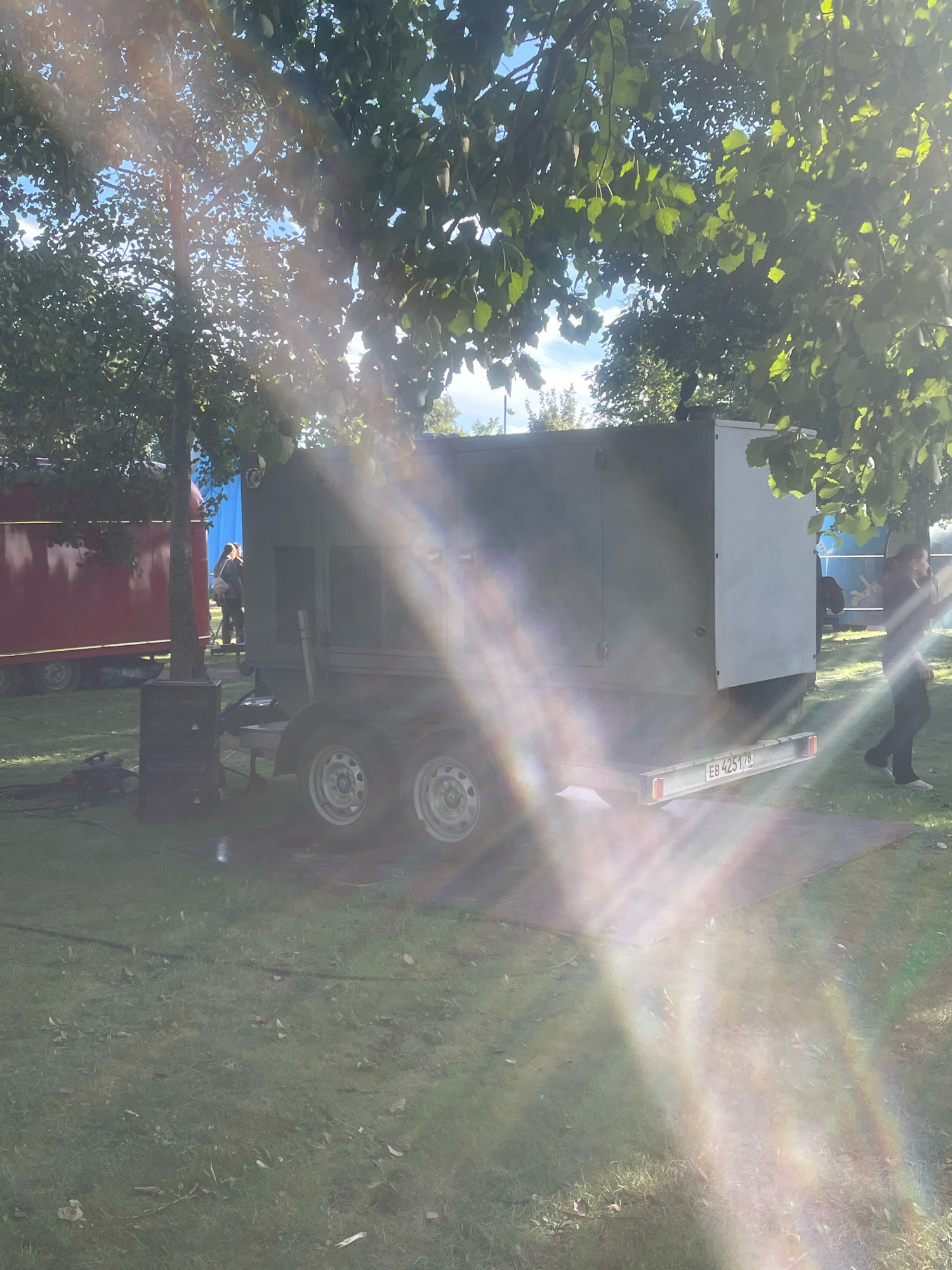 The image shows an outdoor scene, likely a park or festival grounds, with a large generator unit on a trailer in the foreground. The generator is dark gray and has a license plate that reads "EB 425 178". A person is walking away from the generator towards the right of the frame, wearing dark clothing and white shoes. To the left of the generator is a red trailer or structure. The scene is partially obscured by sunlight filtering through the leaves of trees overhead, creating lens flare effects. The ground is covered in grass with some scattered leaves. The overall impression is a casual, outdoor event setup. The image's location is not specified in the Saint Petersburg, Russia context within the image itself.
