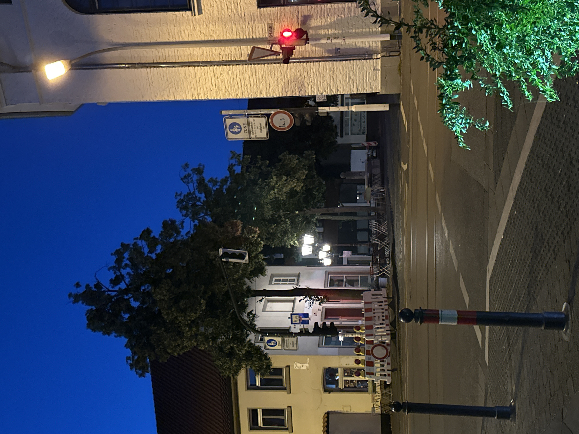 The image depicts an intersection at dusk in Krefeld, Germany. The sky is a deep, clear blue, indicating either early evening or late twilight. A white brick building is prominent on the right side of the frame, with a streetlamp illuminating its facade. A traffic signal with a red light visible is mounted on the building. A street sign indicating "Schulstr." is also present on the building.
On the left side of the intersection, a light-colored building with a dark tiled roof is visible. In front of it, a large tree with dark foliage stands. A traffic light is also present here. A temporary construction barrier, consisting of white and orange fencing with circular red and white signs, blocks a section of the road. Several small round tables and chairs are set up on the sidewalk in front of the building on the left and further down the street on the right, suggesting outdoor seating for a cafe or restaurant.
Two bollards, one dark and one with red and white bands, stand on the sidewalk. The road surface is asphalt, with painted white lines indicating lanes and pedestrian crossings. A patch of lush green foliage is visible in the bottom right corner, illuminated by what appears to be artificial lighting. The scene is quiet, with no people visible. The overall impression is one of a European street scene transitioning into night.