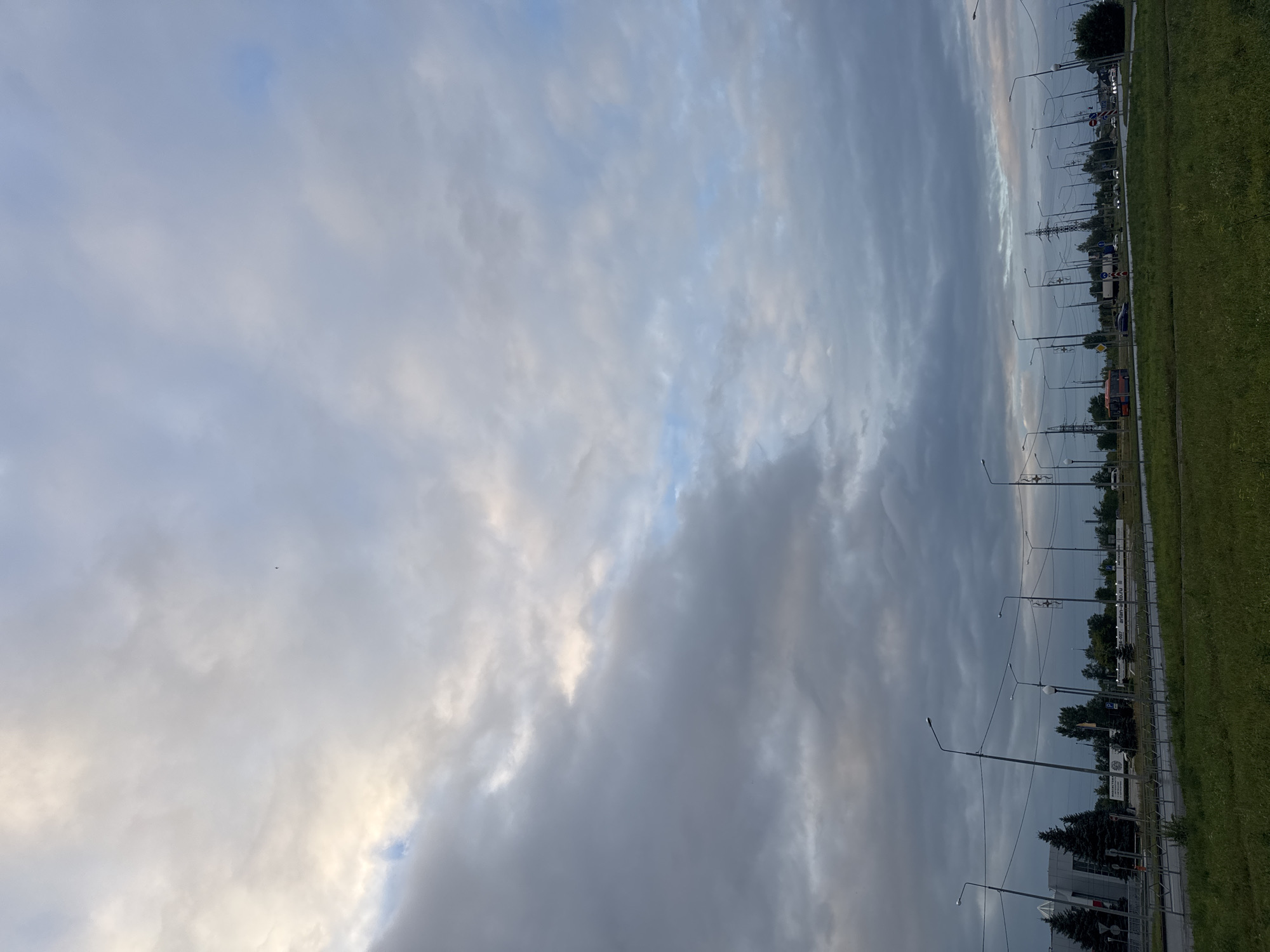 The image shows a view of a cloudy sky over a street with a grass median in Tolyatti, Russia. The sky is filled with a mixture of gray and white clouds, with patches of blue visible. The street appears to be a multi-lane road with traffic lights and streetlights. There are several vehicles on the road, including cars and a bus. In the background, there are buildings and trees. The overall scene suggests a typical urban environment during a cloudy day.