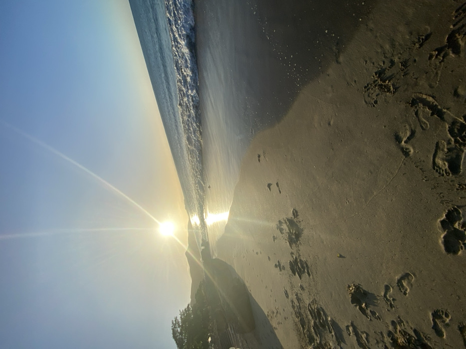 A rotated view of a beach at sunrise or sunset shows the sea's edge meeting a sandy shore. Footprints are imprinted in the sand. A bright sun is positioned on the horizon, casting a long glare across the scene. The sky is a clear gradient of blue. Some trees are visible at the bottom of the scene.