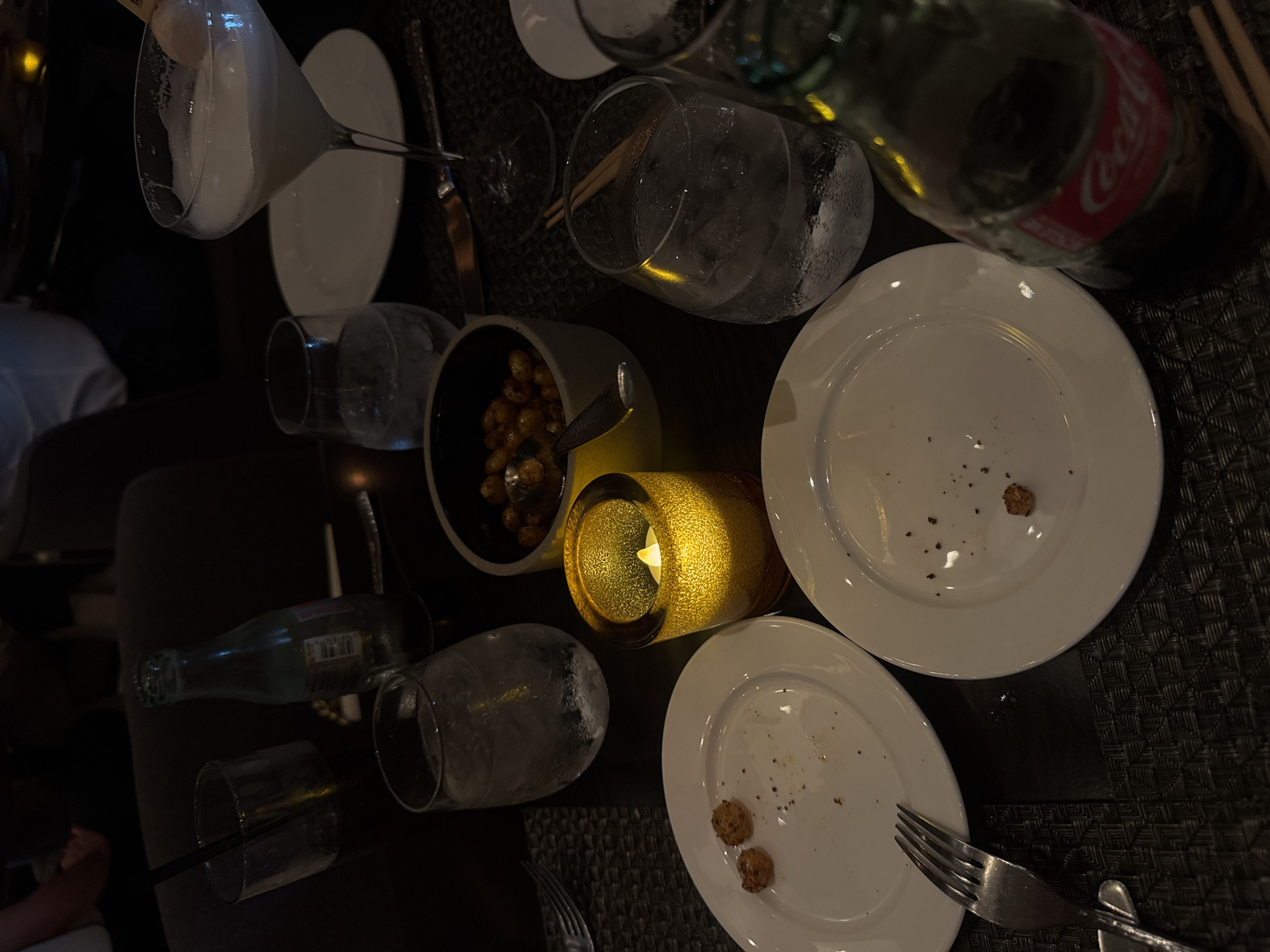 The image shows a dark, intimate restaurant table setting, likely after a meal. The table is covered with a textured, dark grey placemat. There are several plates, some with food remnants, indicating a meal has been consumed. Scattered around the table are various glasses, some containing ice, and a bottle of Coca-Cola. A small bowl contains what appears to be nuts. A lit, decorative candle is placed in the center, providing a warm glow. Forks and knives are positioned near the plates. The setting suggests a sophisticated dining experience. The location in Boca Raton, United States of America, is not identifiable from the image alone.
