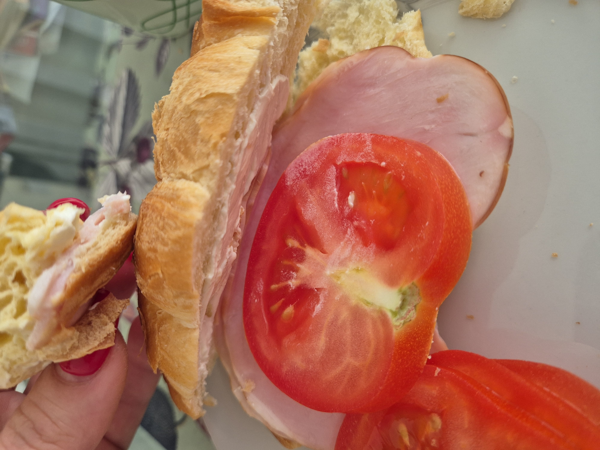 A partially eaten sandwich is displayed on a white plate. The sandwich consists of a sliced croissant roll, ham, and tomato slices. A hand with red nail polish is holding a piece of the croissant. The plate sits on a reflective surface with a floral pattern, suggesting it might be a table. The location is in West Suffolk, United Kingdom.