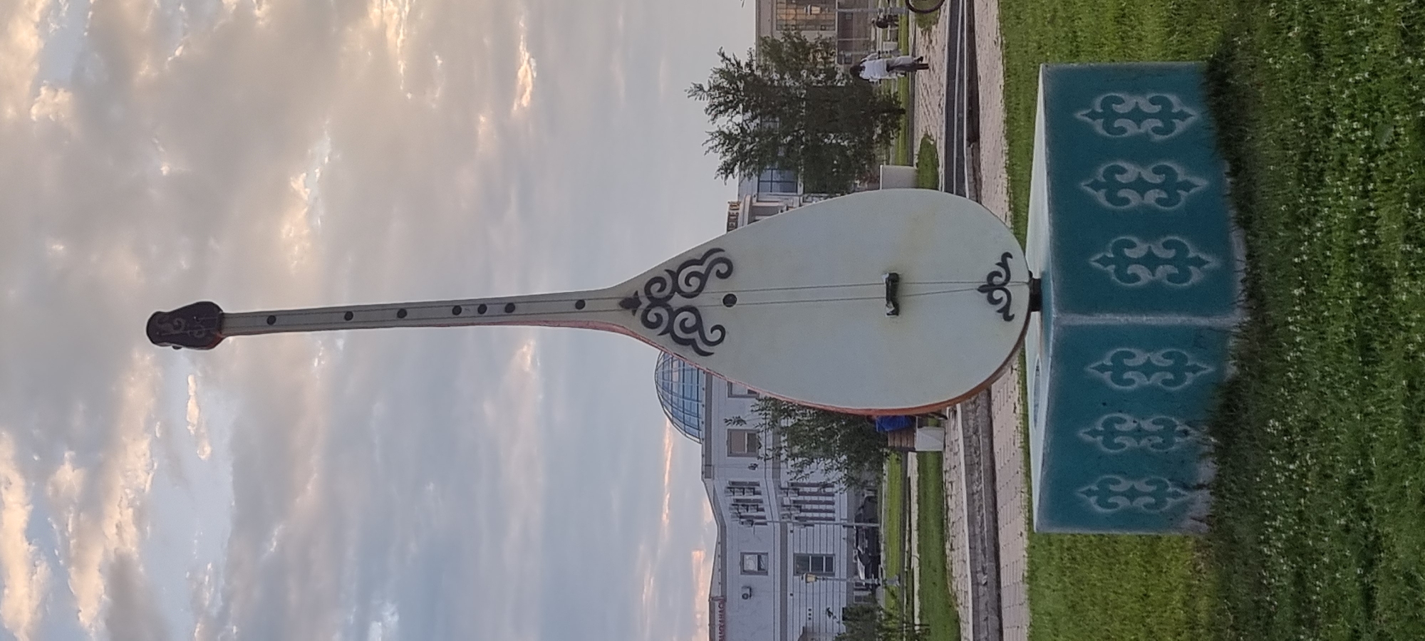 The image shows a large statue of a dombra, a traditional Kazakh string instrument, on a decorative pedestal in a park. The dombra statue is white with black decorative patterns on its body and a dark head. The pedestal is teal with white patterns. In the background, there is a modern building with a dome and trees. There are also people in the background, including one person riding a bicycle. The sky is cloudy with hints of sunset colors. This statue is located in the central park of the city of Aktobe, Kazakhstan.