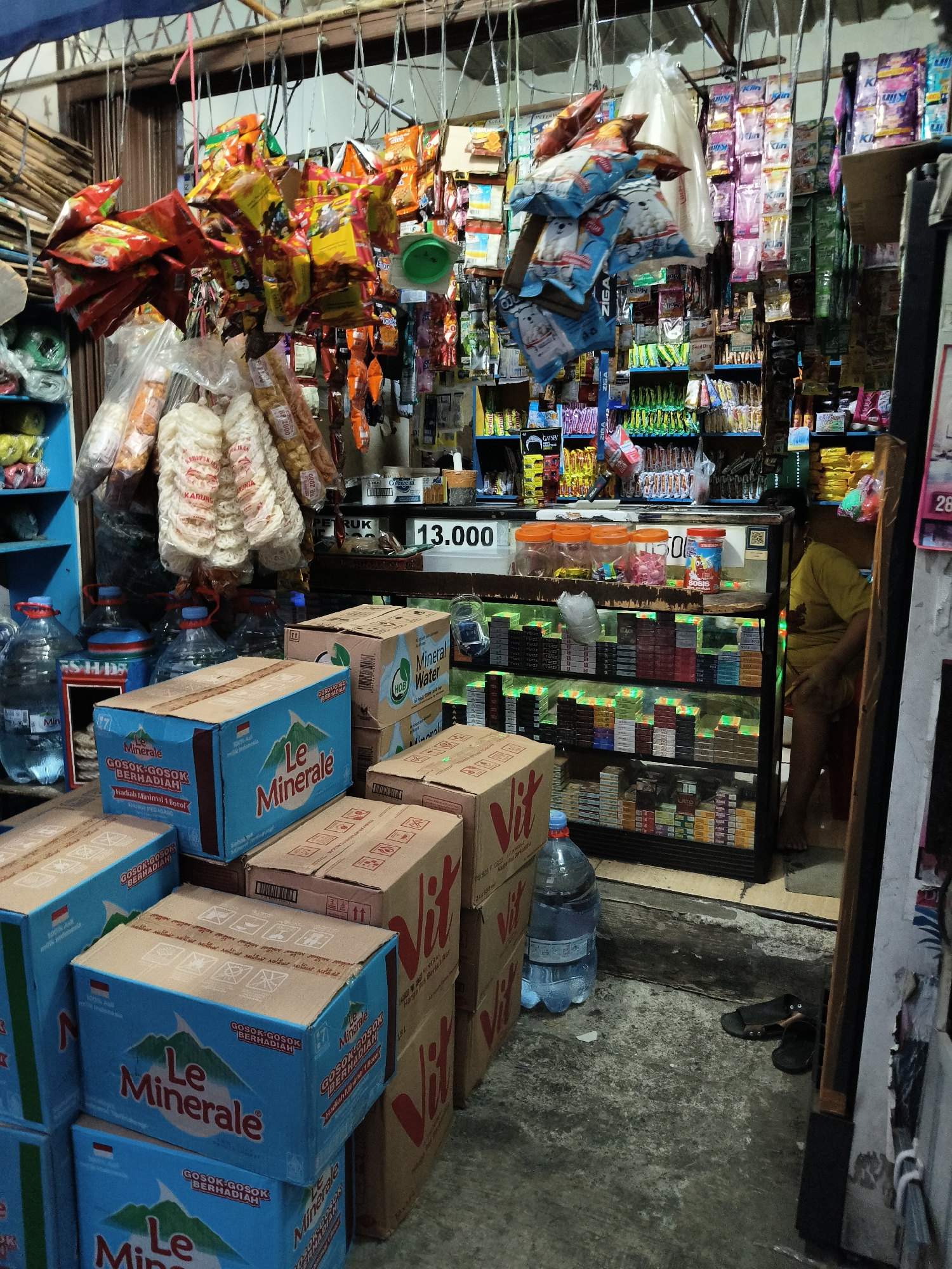 This is an image of a small convenience store, likely a warung, located in South Tangerang, Indonesia. The store is densely packed with various goods, including packaged snacks, drinks, and cigarettes, predominantly displayed on shelves and hanging from the ceiling. In the foreground, stacks of blue cardboard boxes labeled "Le Minerale" and "Vit" are visible, suggesting bottled water or other beverages are being sold. Large plastic bottles of water are also present. A glass display case filled with cigarette packs is prominently featured in the middle ground. A person in a yellow shirt is partially visible in the background, appearing to be the shopkeeper or an employee. The floor is concrete and somewhat worn. The overall impression is of a bustling, local retail establishment typical of Indonesian neighborhoods.