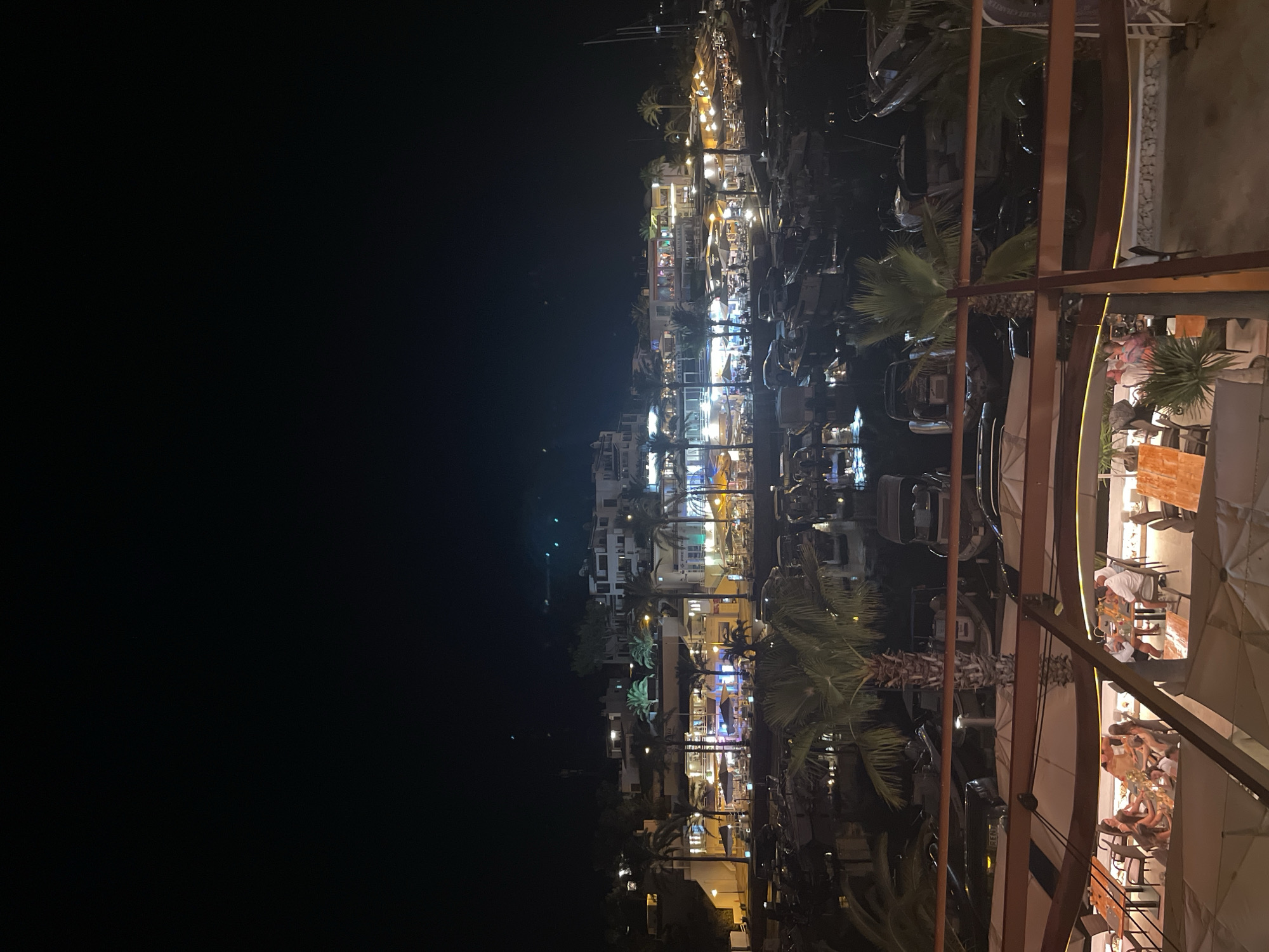 The image depicts a nighttime scene at a marina in Santanyí, Spain, specifically the marina of Cala d'Or. The foreground shows an outdoor dining area with tables and chairs, some occupied by people. The lighting is warm and atmospheric, with umbrellas providing shade over the seating areas. In the background, a row of buildings with lit windows and balconies lines the waterfront. Numerous boats are docked in the marina, their reflections shimmering on the dark water. Palm trees are scattered throughout the scene, adding to the Mediterranean ambiance. The sky is dark, indicating it is nighttime, with a few faint lights visible in the distance.