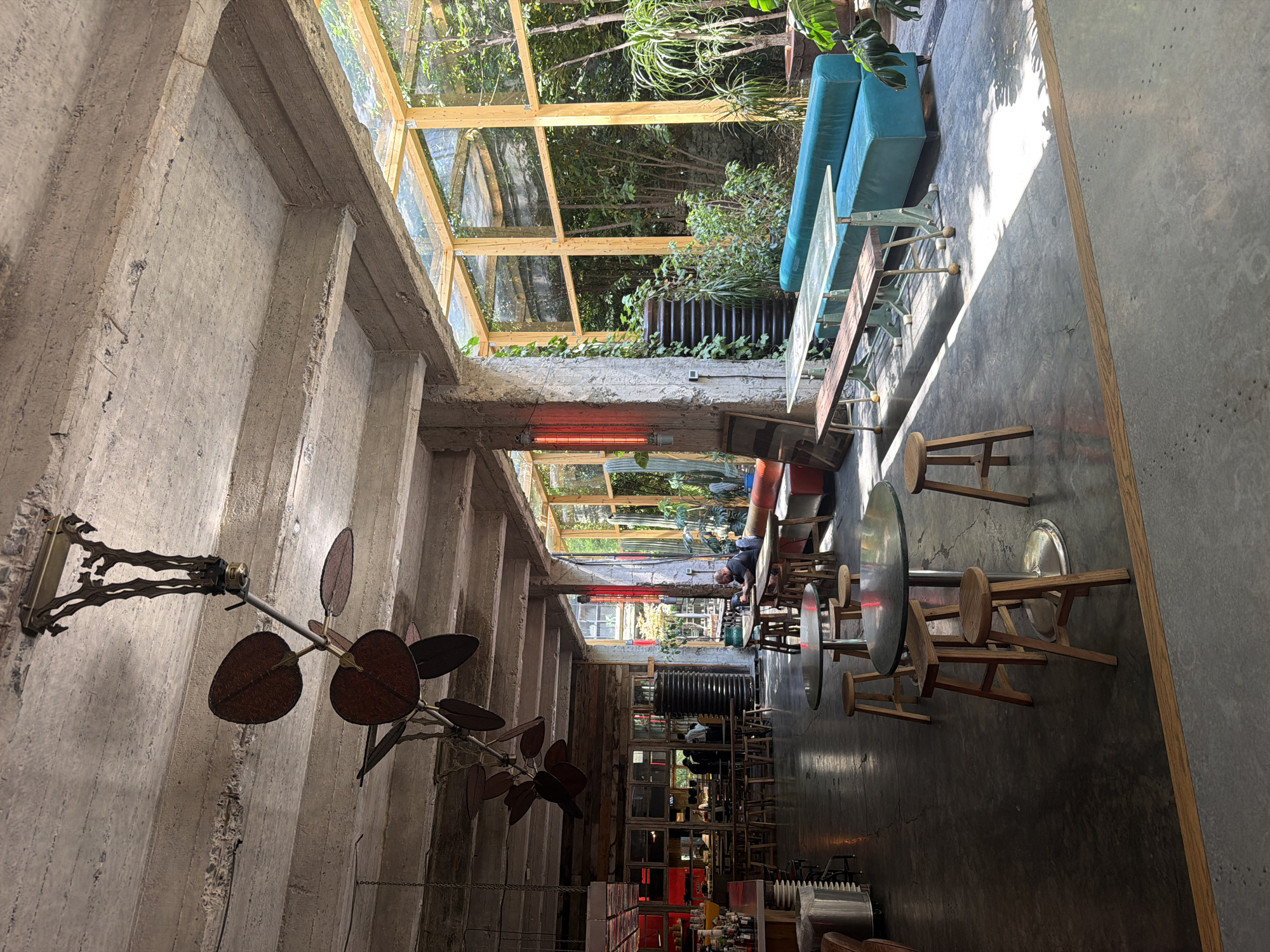 The image depicts the interior of a restaurant with an industrial-chic aesthetic, likely located in Tbilisi, Georgia. The space features exposed concrete ceilings and pillars, with wooden beams and a glass-enclosed garden area visible. Sunlight streams into the room, casting long shadows on the concrete floor.
In the foreground, several round tables with wooden stools are arranged. Further back, a long bench seat upholstered in a vibrant teal fabric is paired with a wooden bench and a glass-topped table in the sunlit garden area.
The background reveals a bar area with shelves stocked with bottles and glasses. Several people are present, including a man sitting alone at a table in the garden area, and a few individuals are visible behind the bar. A distinctive ceiling fan with large, dark blades is suspended from the concrete ceiling. A red heating element is mounted on a pillar near the garden entrance. The overall impression is a spacious and somewhat rustic dining environment with an outdoor ambiance.