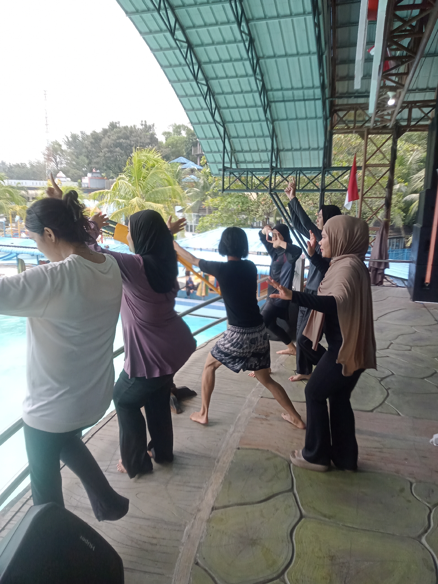 In Baru, Indonesia, several individuals are engaged in what appears to be a dance or synchronized movement routine near a swimming pool. The setting is outdoors, under a large, green-slatted canopy. The group consists of at least six people, all facing towards the pool or slightly away from the camera.
The person closest to the camera on the left is wearing a white long-sleeved shirt and dark pants. To their right, another person in a purple long-sleeved shirt and dark pants is facing away from the camera with arms extended outwards. A third person, also facing away, wears a black short-sleeved shirt with patterned shorts and dark pants. Further to the right, a person in a black long-sleeved shirt and dark pants has their right arm raised. Next to them, a person wearing a beige headscarf and black clothing is in a similar pose with arms outstretched. Another individual in black clothing is also visible, partially obscured.
The environment includes the swimming pool on the left, with blue water and railings. Beyond the pool, palm trees and other foliage are visible, suggesting a tropical or sub-tropical climate. In the background, a tall antenna structure can be seen. The ground where the individuals are standing is a textured, stone-like surface in green and beige tones. A black speaker is positioned in the foreground on the left. Red and white Indonesian flags are hung from the canopy structure. The overall impression is of an organized activity taking place in a recreational area.
