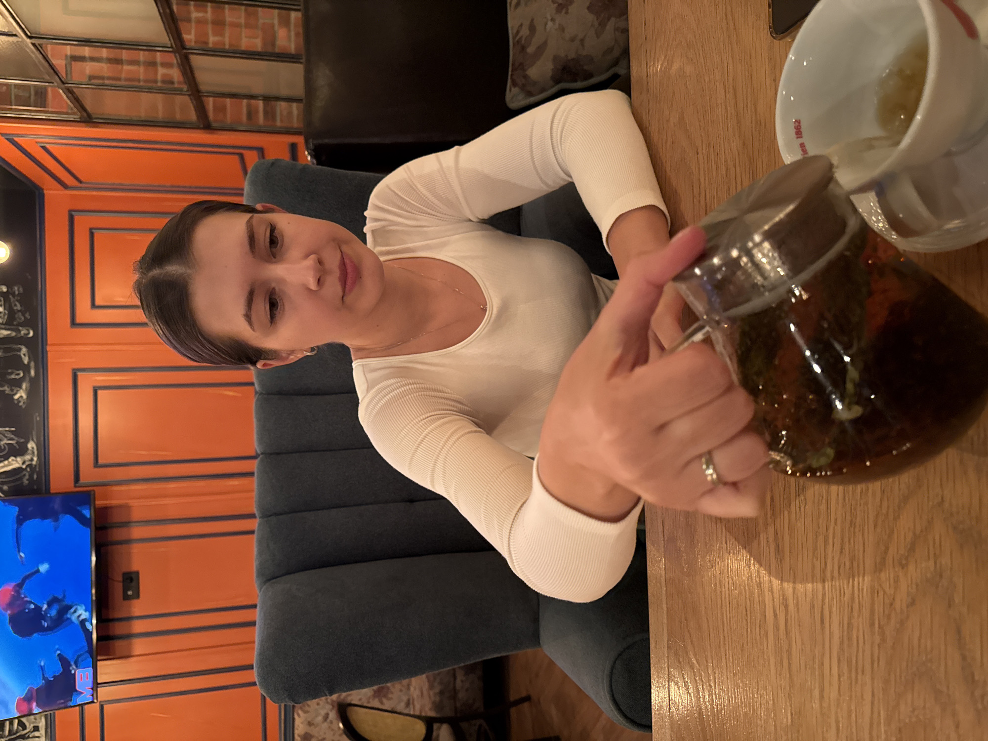 The image shows a woman seated at a wooden table, pouring tea from a glass teapot into a white porcelain cup. She is wearing a white long-sleeved shirt and a ring on her right hand. She appears to be in a restaurant, likely located in Krasnoyarsk, Russia based on the writing on the cup. The background features an orange and navy blue paneled wall, a blue tufted chair, and a television screen showing images.