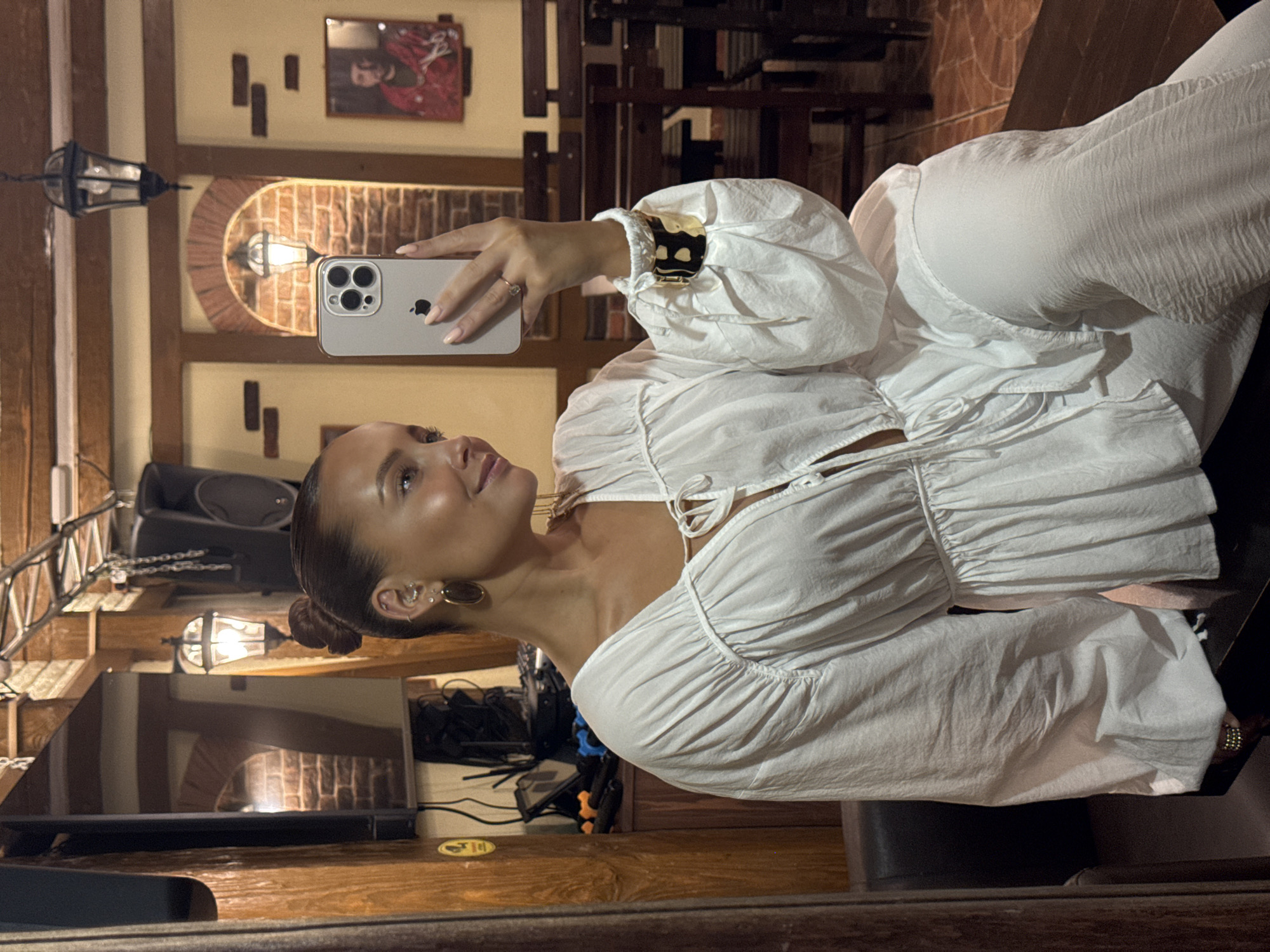 A woman is taking a mirror selfie in what appears to be a rustic establishment, possibly a restaurant or bar. She is wearing a flowing white top with voluminous sleeves and white pants. She has her hair pulled up in a bun and is wearing earrings and bracelets. The background shows wooden beams, exposed brick, and hanging lamps, consistent with a traditional or themed interior. A framed picture of a man in a red garment is visible on the wall. The lighting is warm and dim. The image does not contain any information indicating the location as Chaikovsky, Russia.