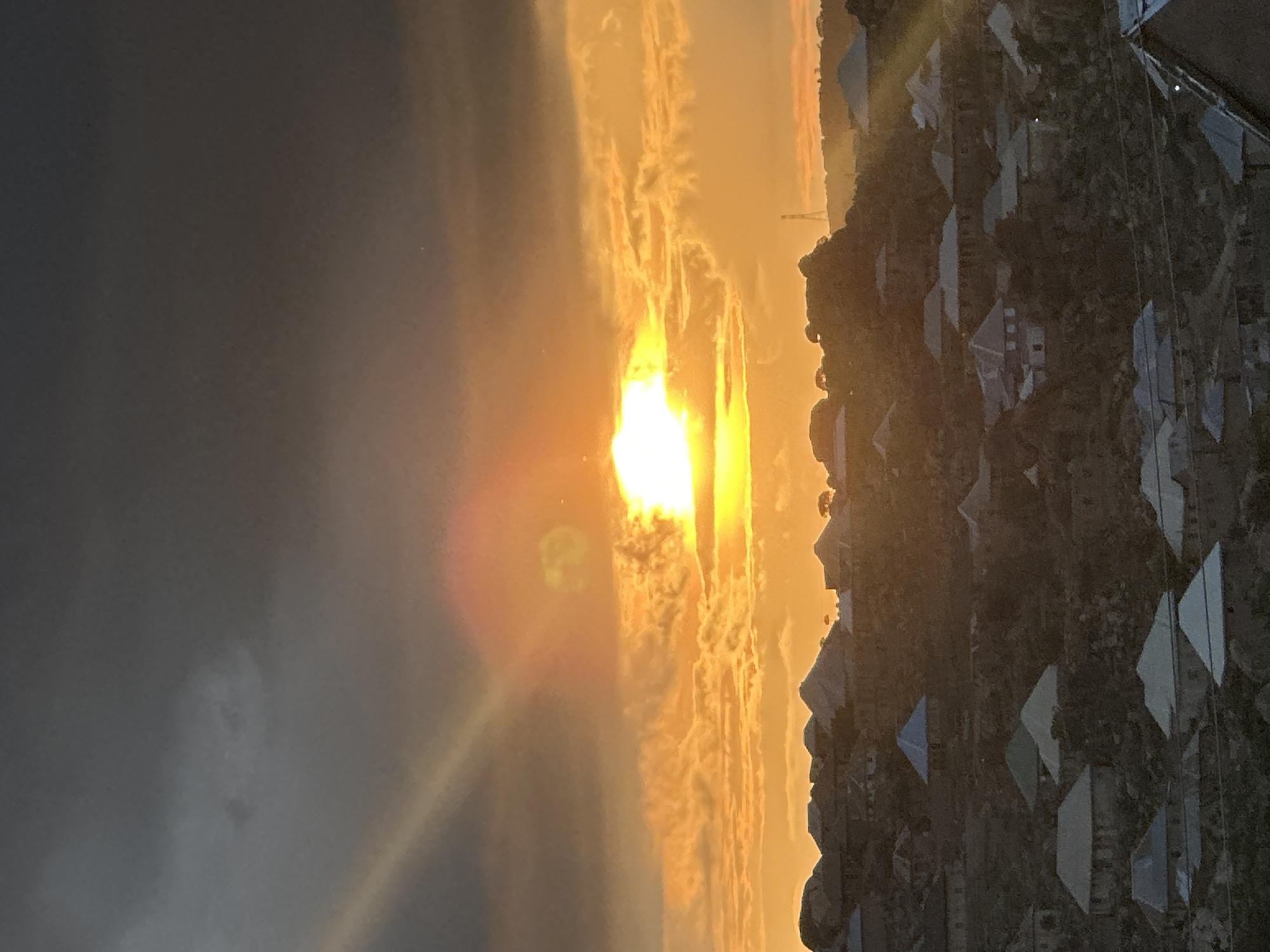 The image captures a sunset or sunrise over a densely populated, hilly residential area within the city of Jos, Nigeria. The sky occupies the upper portion, displaying dark grey stratocumulus clouds in the zenith, transitioning to vibrant orange and yellow hues near the horizon where the sun is positioned. The sun, a brilliant white-yellow orb, is partially obscured by clouds, emitting strong lens flare and rays of light that extend diagonally across the frame.
Below the sky, the landscape consists of undulating hills covered extensively with residential structures. These houses exhibit varied architectural styles, featuring a mix of conical, gabled, and flat roofs, predominantly in shades of grey, blue, and brown. The terrain appears rugged and rocky, with sparse vegetation interspersed among the dwellings. No individuals are visible, and no specific actions or interactions are discernible. In the far background, positioned on a distant hill, a tall, slender communication tower or similar mast is identifiable against the bright horizon.