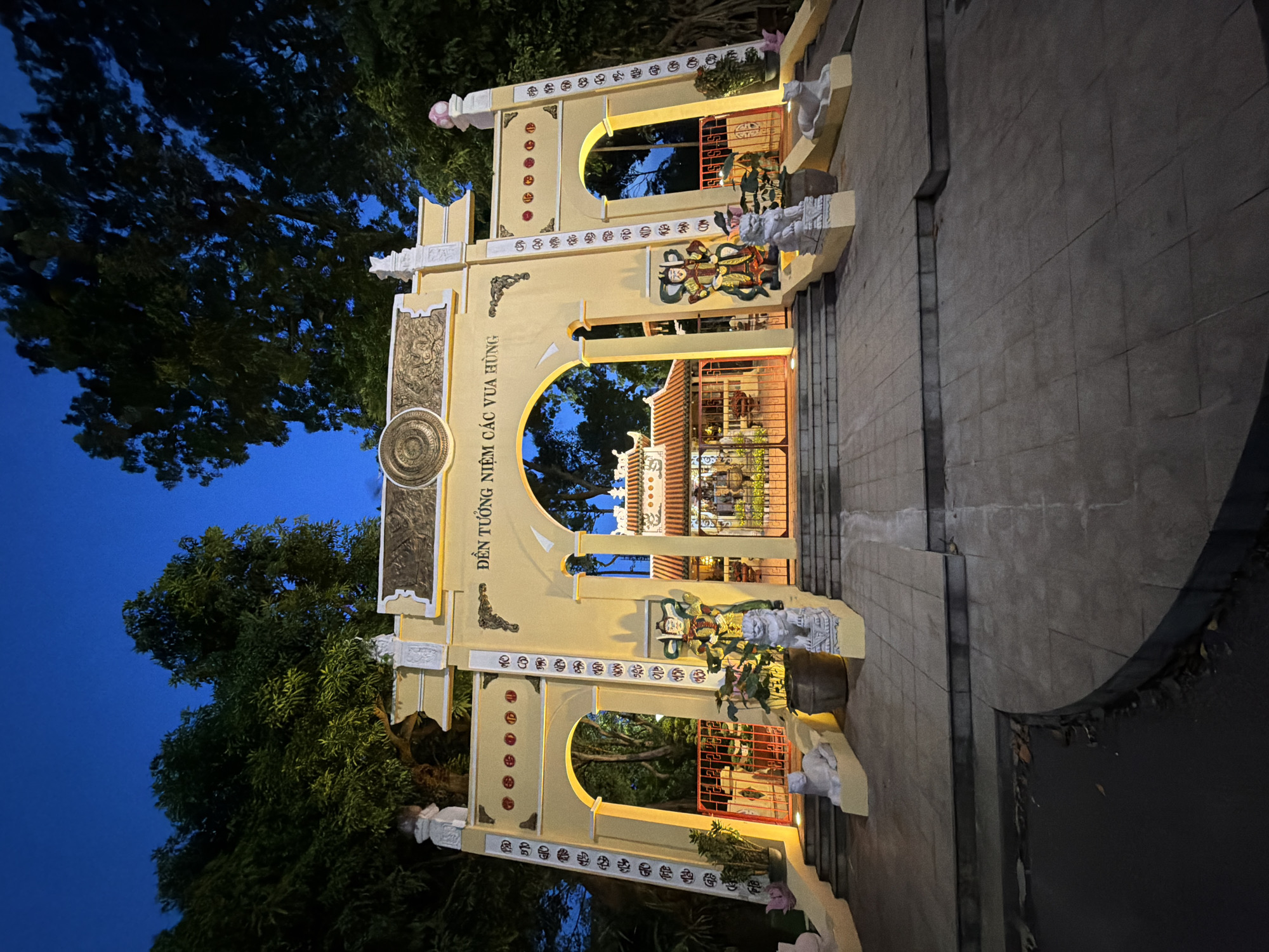 This is a vertical, eye-level, medium shot of the yellow archway of the Temple of the Hung Kings in Thủ Đức City, Vietnam, at dusk. The archway is ornate, with decorative carvings and script. The entrance to the temple is visible through the central arch, featuring traditional Vietnamese architecture with a tiled roof. Two guardian statues, resembling warriors and lions, flank the entrance. The area is illuminated by yellow lights, highlighting the yellow paint of the arch and casting shadows. Surrounding the temple are lush green trees, their dark silhouettes contrasting with the bright blue twilight sky. The foreground shows a paved walkway with steps leading up to the archway.